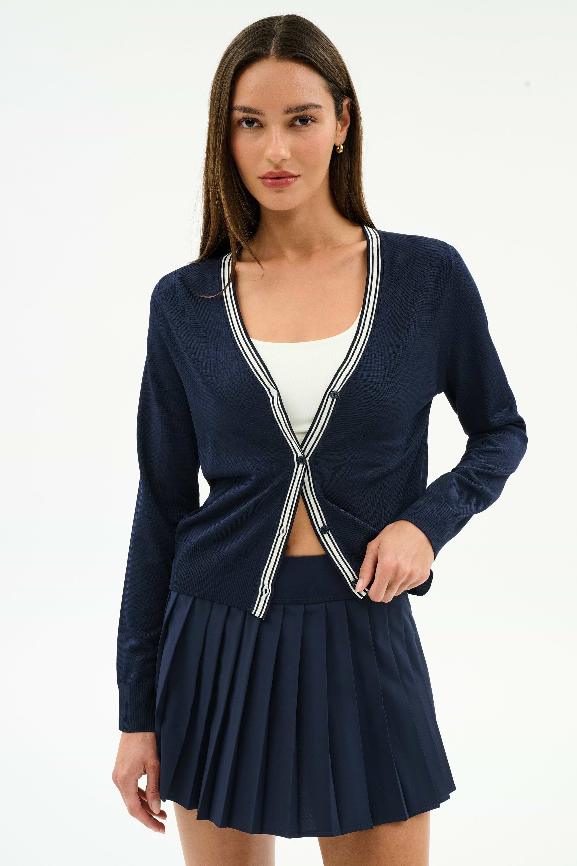 A woman wears the SPLITS59 Bekah Knit Cardigan in Indigo/White over a white top and a navy pleated skirt, standing against a simple light background.