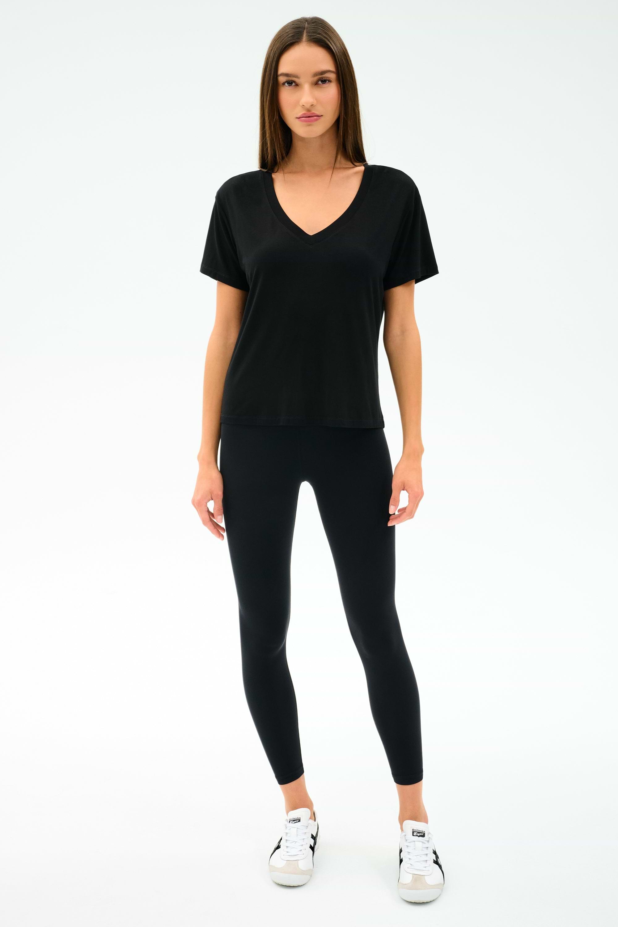 A woman faces forward, wearing the SPLITS59 Georgia Jersey V-Neck in black—crafted from soft viscose and spandex—with black leggings and white sneakers, set against a plain white background.
