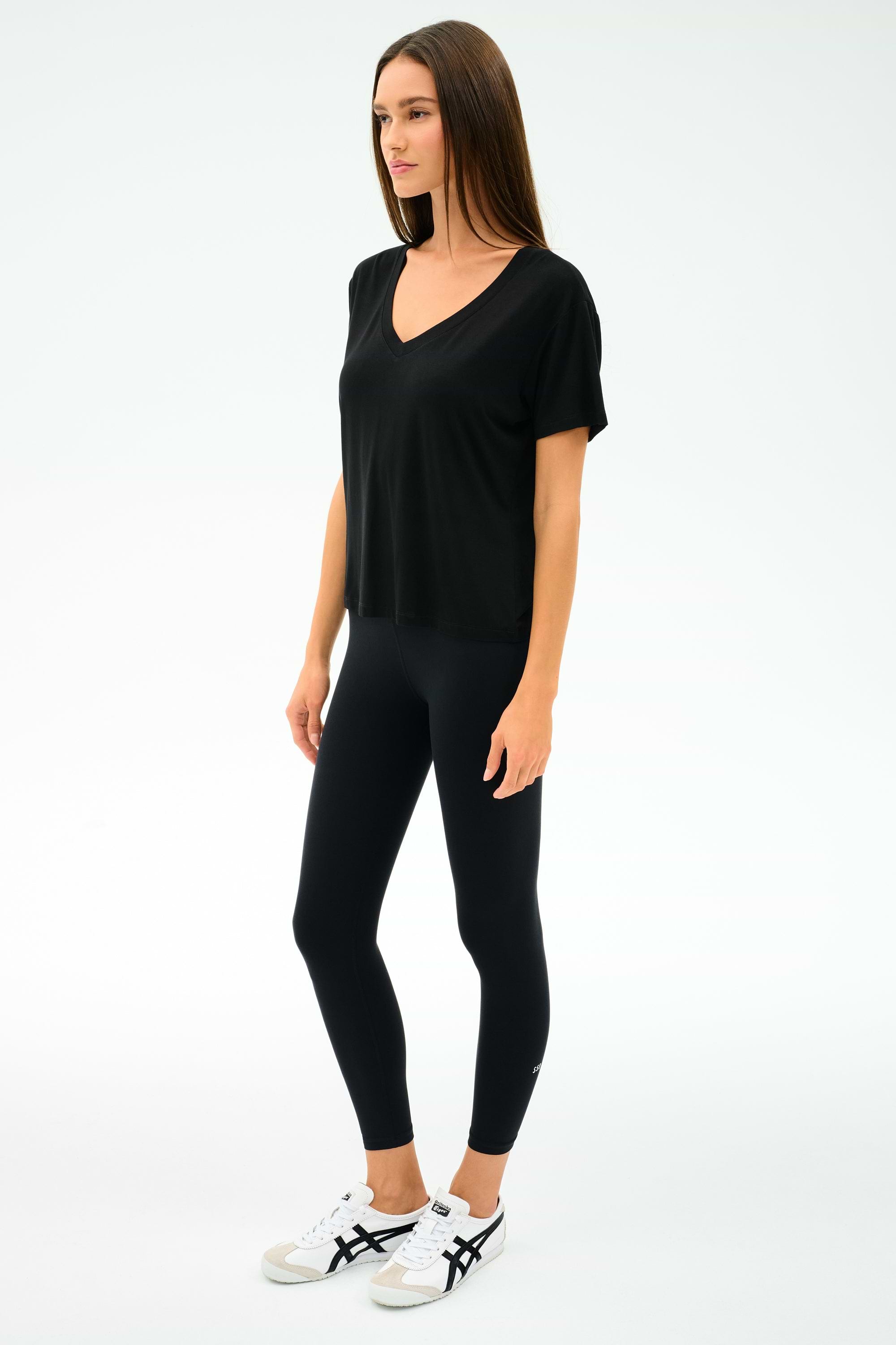 A woman stands against a plain background wearing the SPLITS59 Georgia Jersey V-Neck in black, paired with black leggings and white sneakers with black accents.