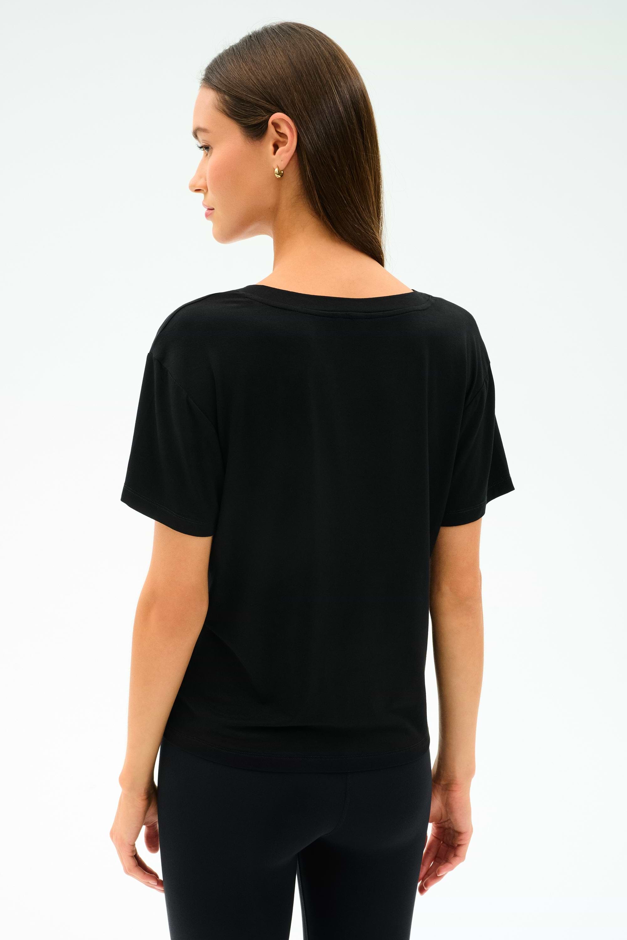 A woman with straight brown hair is standing and facing away from the camera against a plain light background, wearing the SPLITS59 Georgia Jersey V-Neck - Black and black pants.
