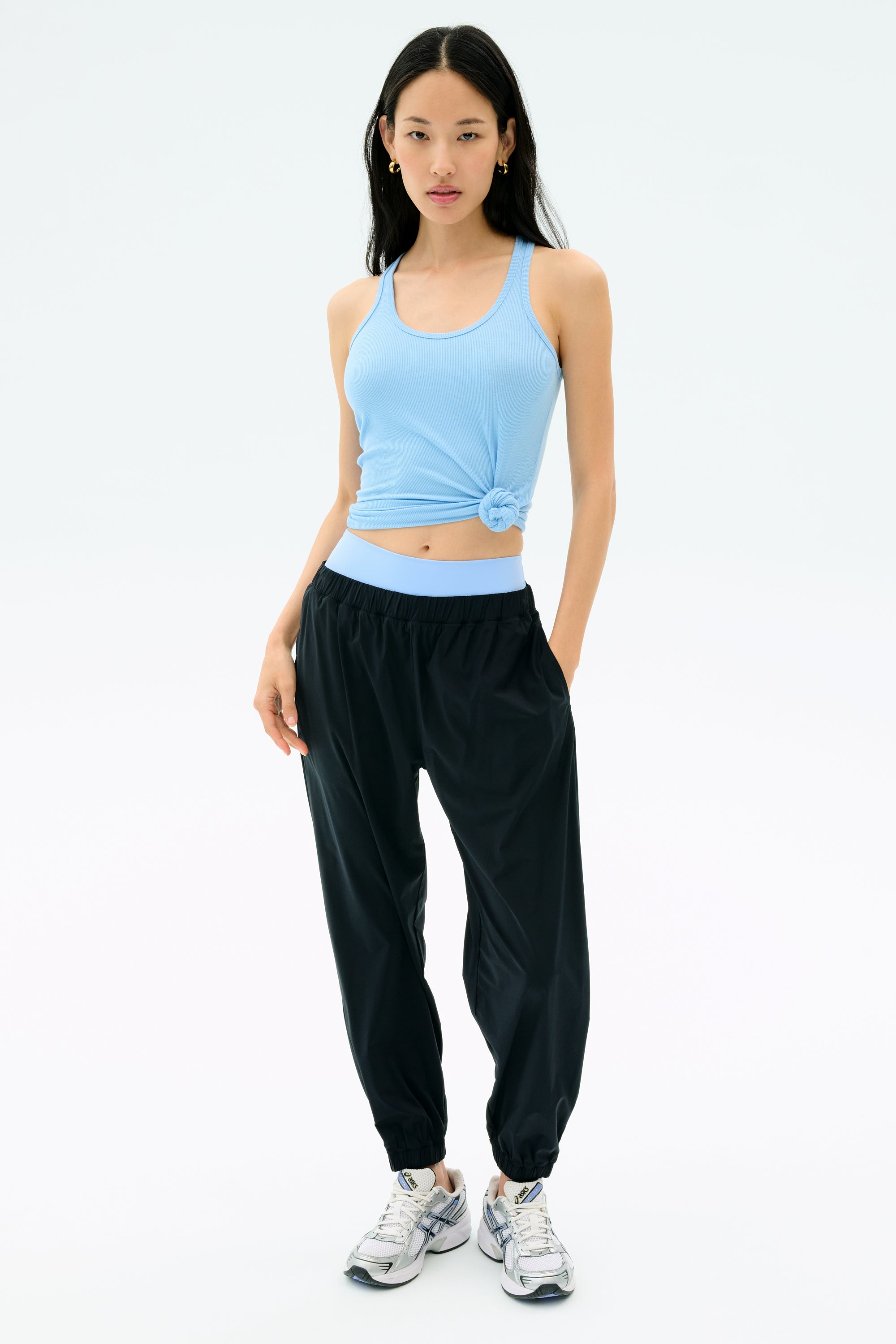 A woman stands against a plain background wearing a light blue spandex tank top, white sneakers, and the SPLITS59 Megan Woven Nylon Jogger in black, with one hand in her pocket.