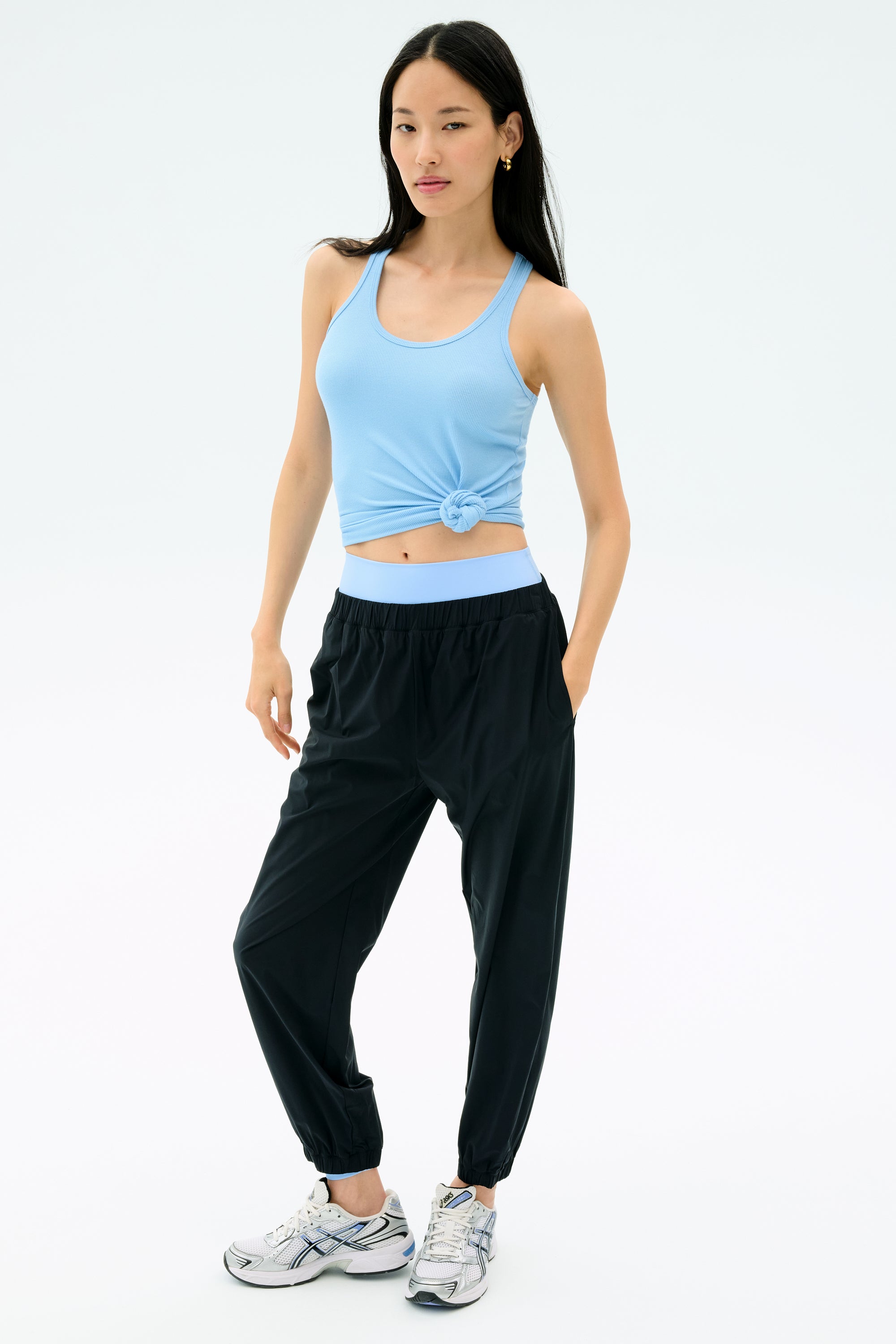 A woman stands against a light background wearing SPLITS59's Megan Woven Nylon Jogger in black, paired with a light blue spandex tank top and white sneakers, her left hand resting in her pocket.