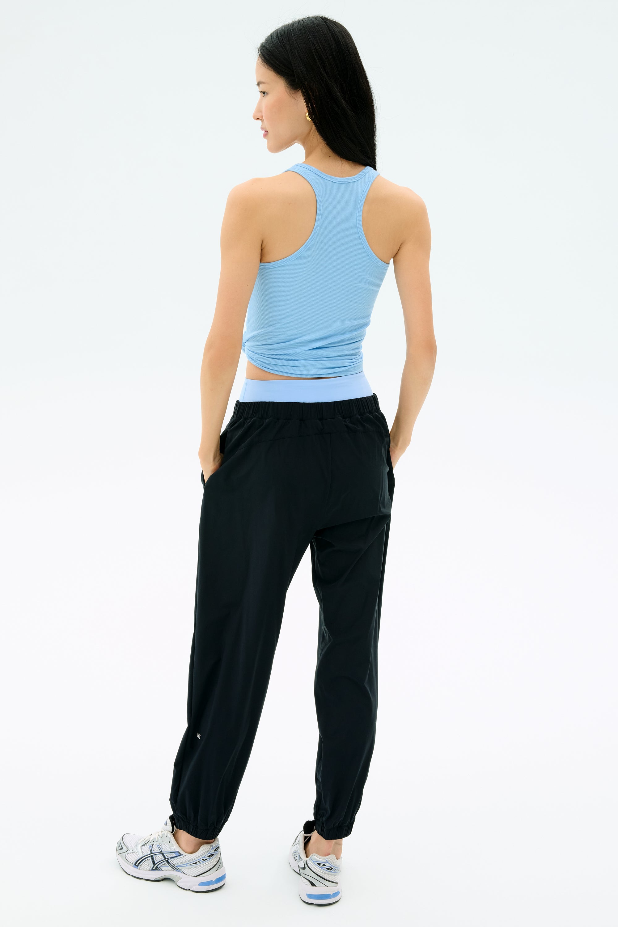 A person faces away, wearing a light blue racerback tank, SPLITS59 Megan Woven Nylon Jogger in black, and white athletic shoes against a plain, light background.