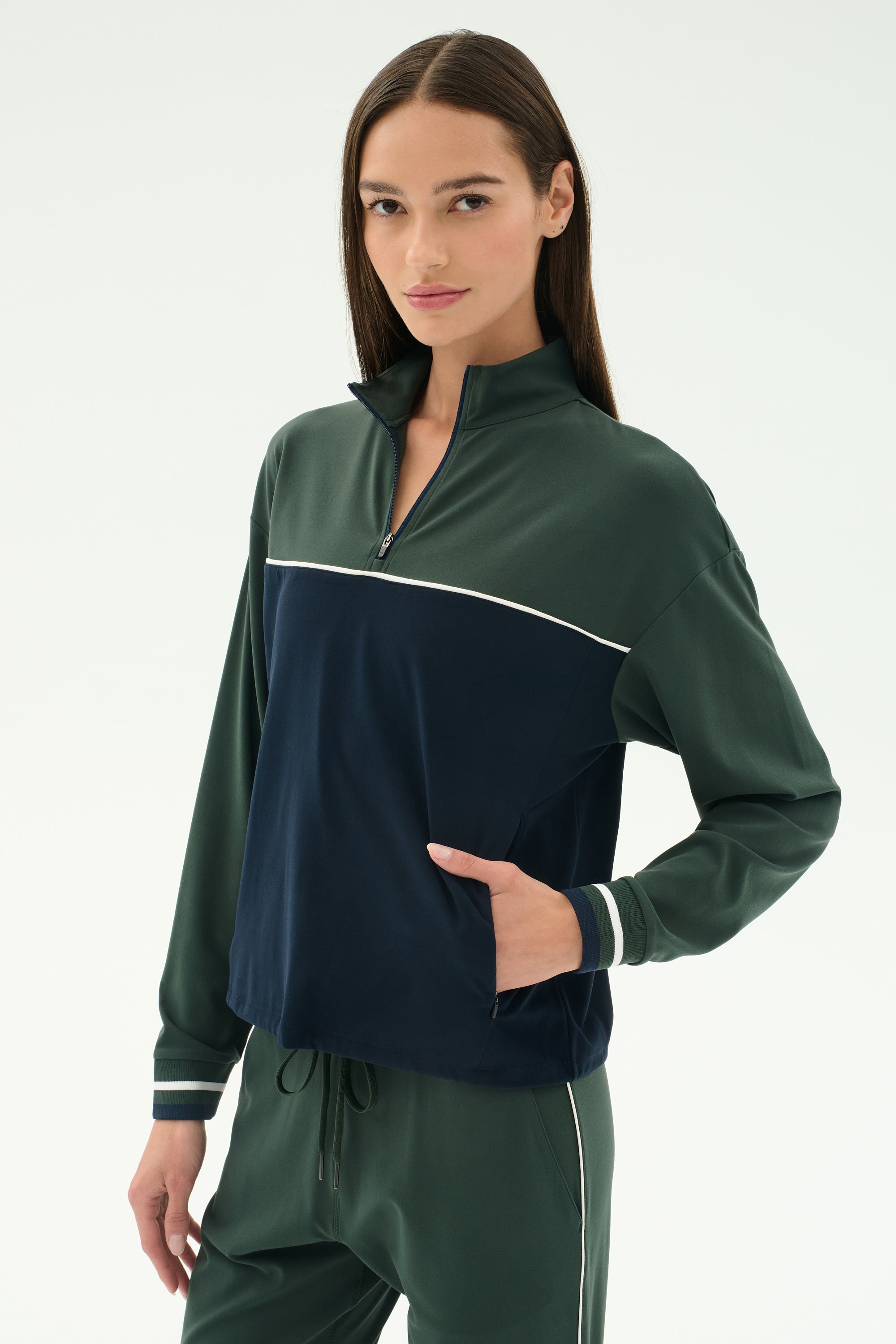 A woman stands against a plain background wearing the SPLITS59 Millie Airweight Half Zip in Military/Indigo with matching pants, featuring white piping and crafted from smooth nylon, with one hand casually placed in her pocket.