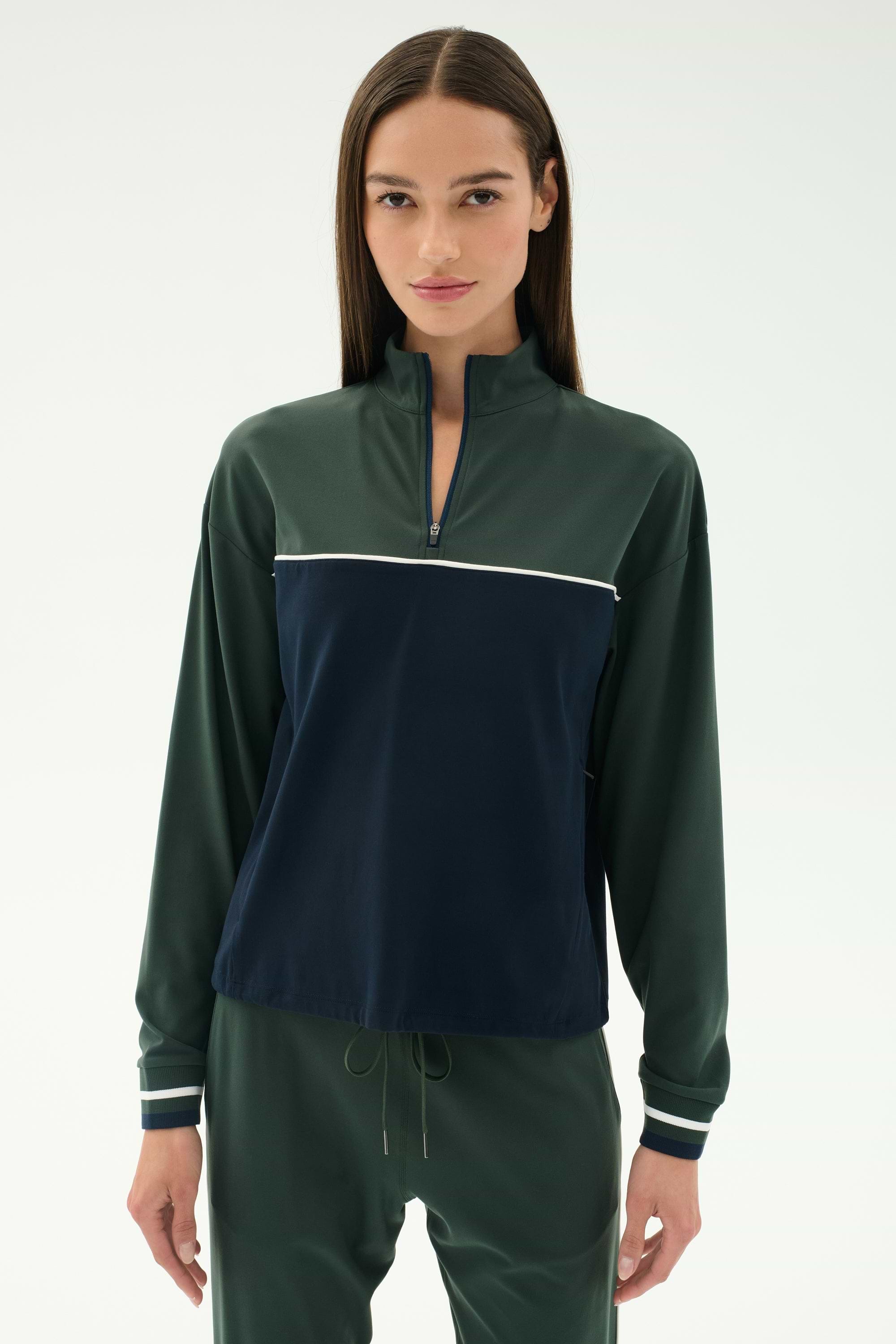 A woman with straight brown hair wears the SPLITS59 Millie Airweight Half Zip in green and navy with matching green pants, standing against a plain light background.