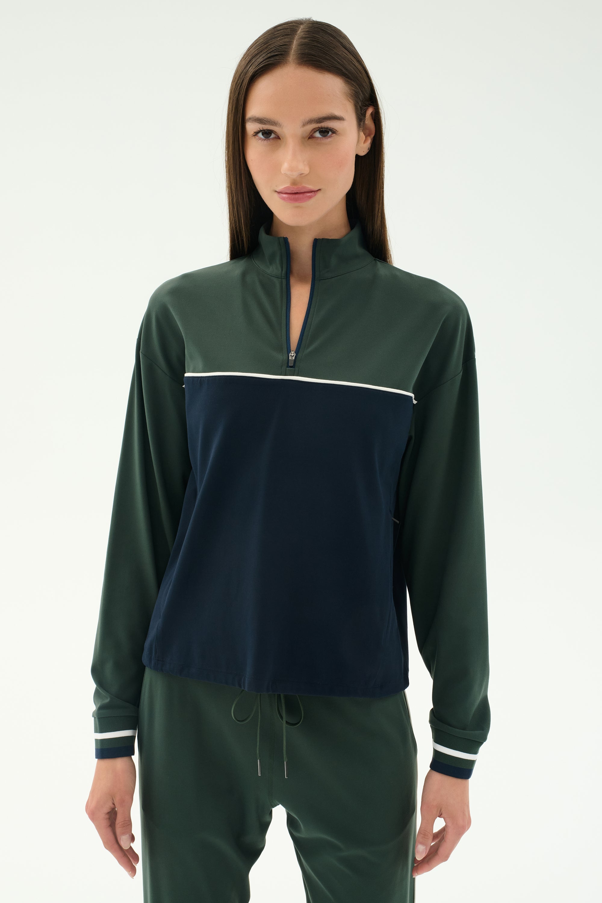A woman with straight brown hair wears the SPLITS59 Millie Airweight Half Zip in green and navy with matching green pants, standing against a plain light background.
