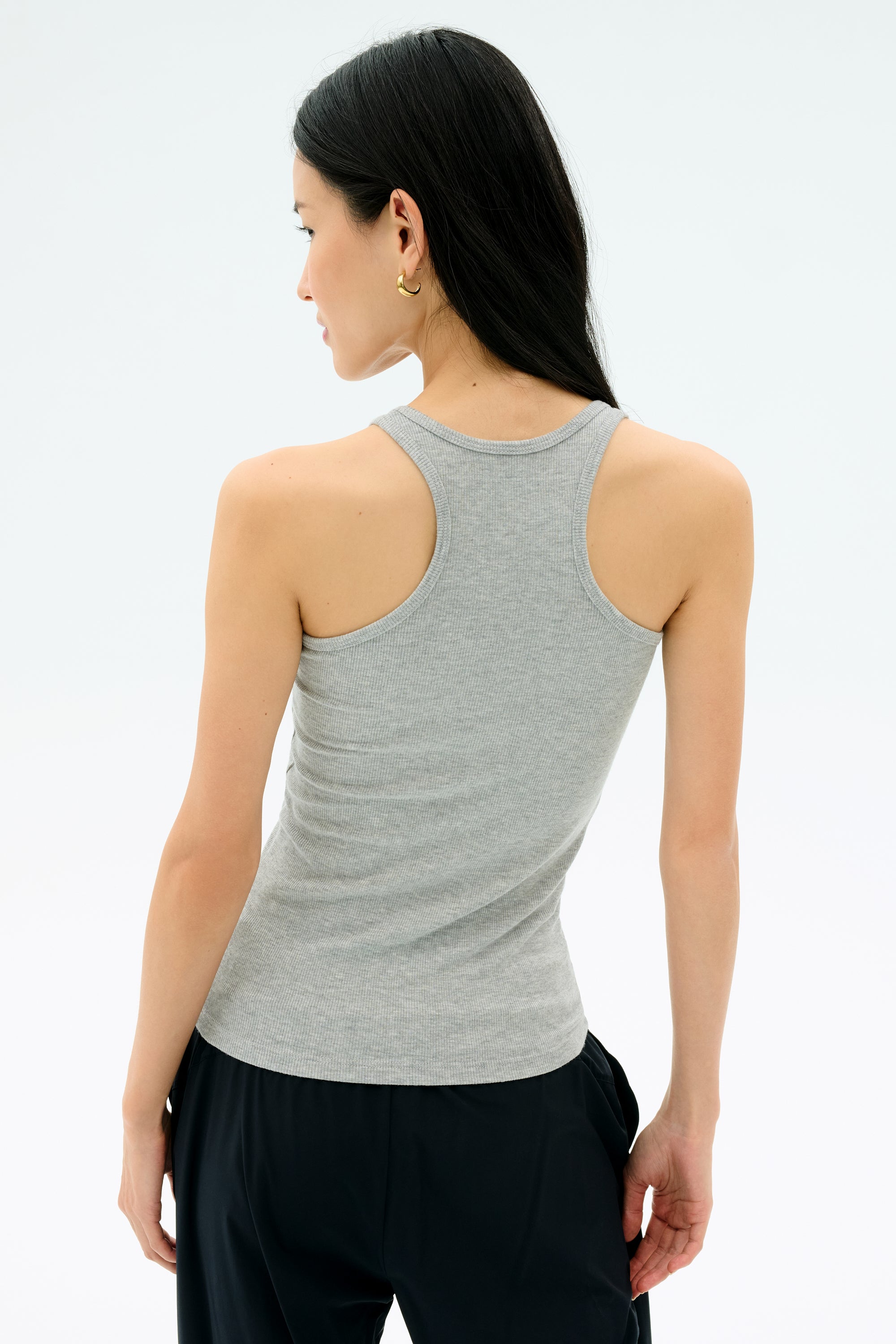 A woman with long dark hair, wearing the Splits59 Ashby Rib Tank in Lt. Heather Grey and black pants, stands facing away against a plain light background.