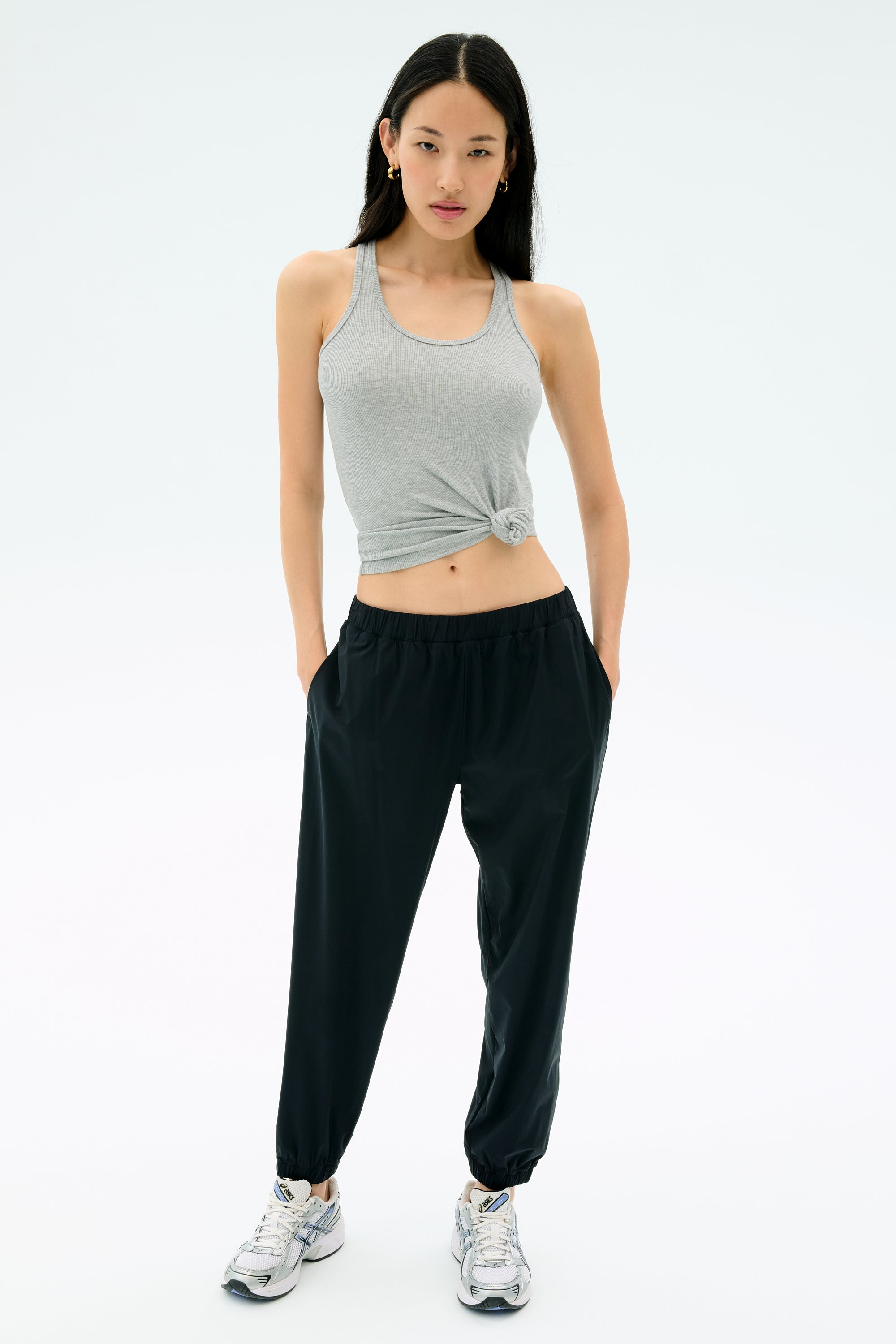A woman faces forward against a plain background, wearing the Splits59 Ashby Rib Tank in Lt. Heather Grey, paired with black jogger pants and white sneakers, with the tank knotted at her waist.