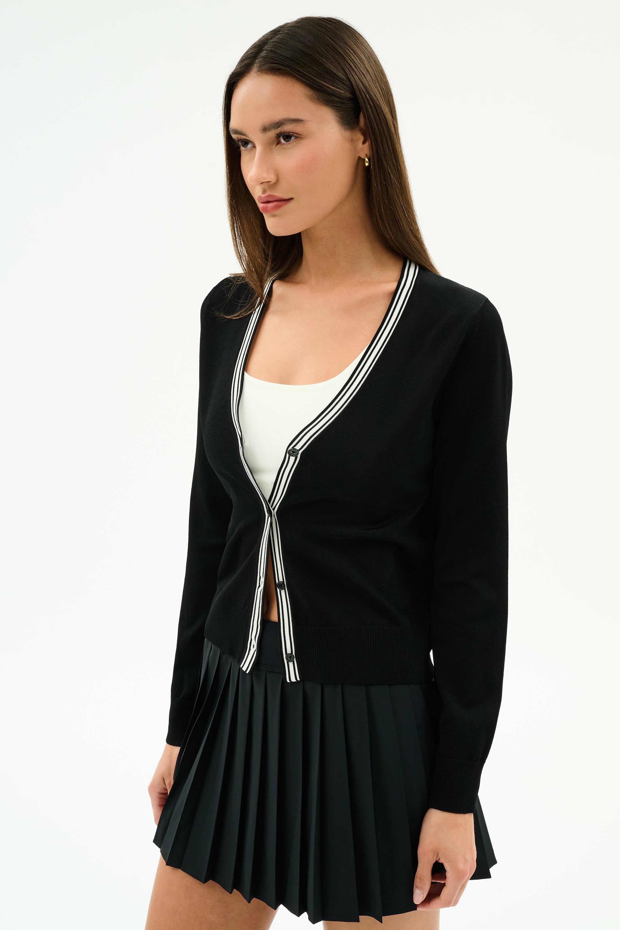 A woman in the SPLITS59 Bekah Knit Cardigan - Black/White over a white top and black pleated skirt stands against a plain white background.