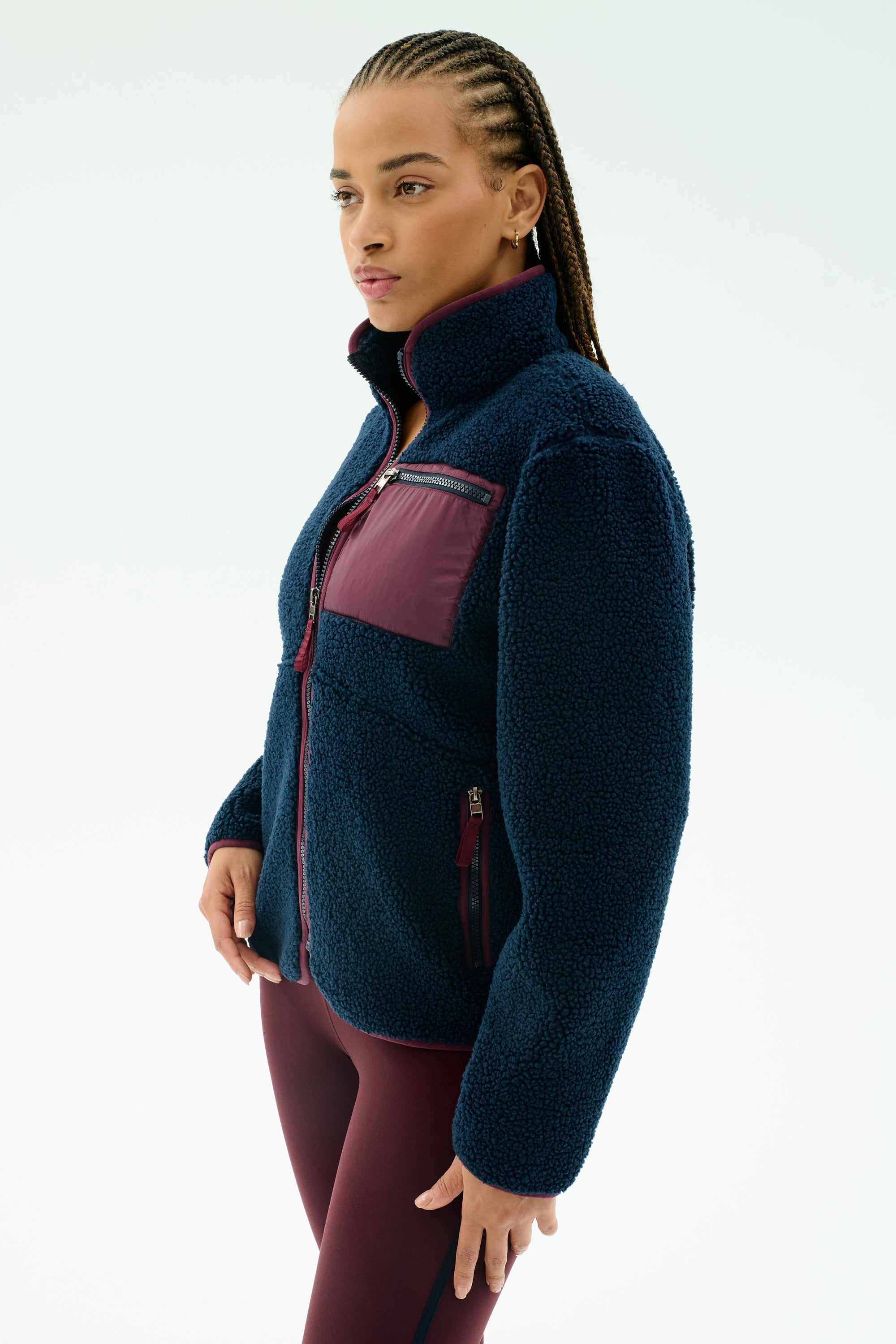 A woman stands against a plain background wearing the SPLITS59 Harris Sherpa Jacket in Indigo/Dark Cherry, featuring soft 100% polyester fleece with a maroon chest pocket and zipper, paired with maroon leggings.