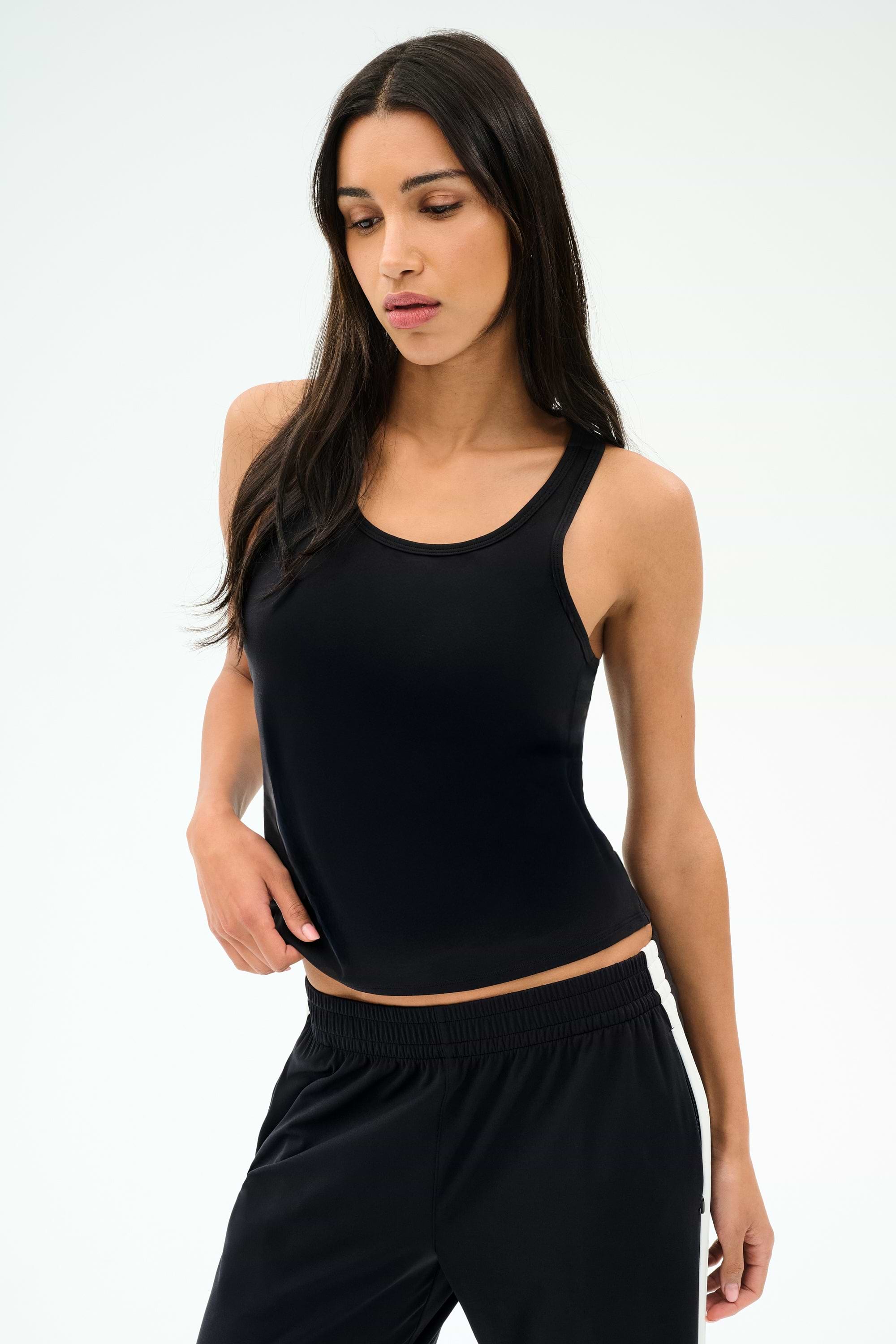 A woman with long dark hair wears the SPLITS59 Mel Airweight Lite Tank in black and black pants, standing against a plain light background.