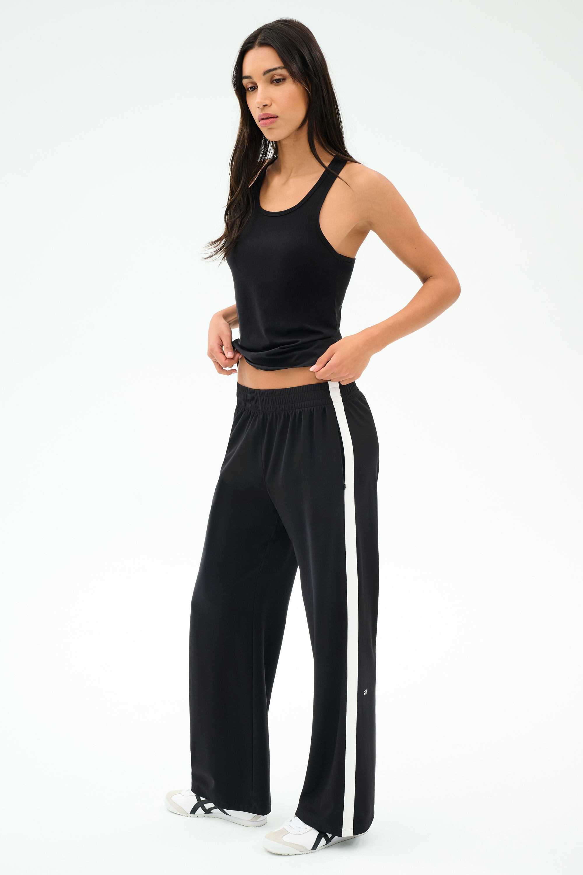 A woman stands against a plain background wearing the SPLITS59 Mel Airweight Lite Tank in black, paired with black wide-leg pants featuring a white side stripe, and white sneakers.