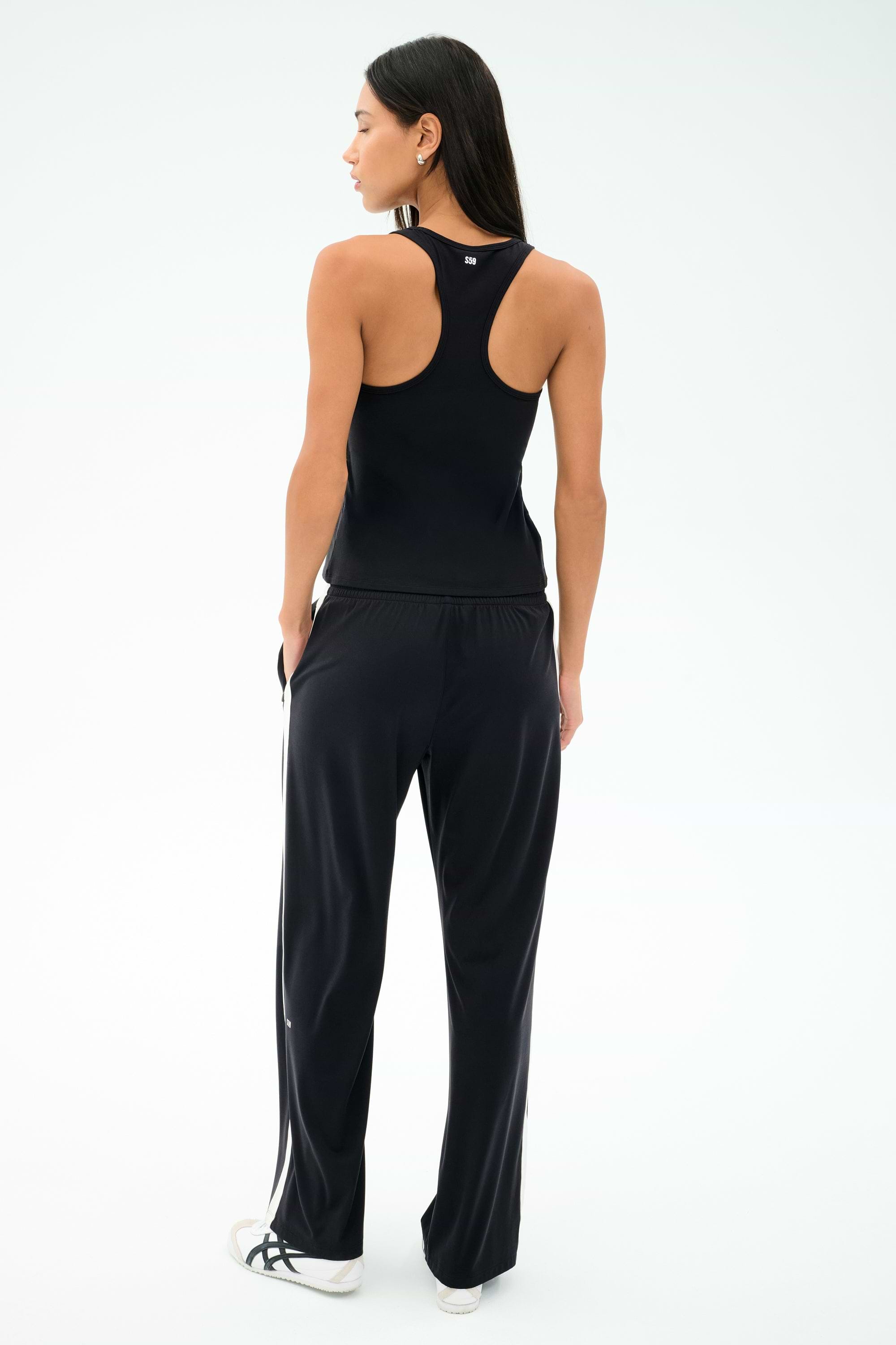 A woman faces away, wearing the SPLITS59 Mel Airweight Lite Tank in black, paired with black track pants with white side stripes and white sneakers, set against a plain light background.
