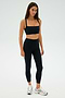 Amidst a minimalist setting, an individual stands confidently in a sleek black SPLITS59 Monah Rigor Bra paired with matching leggings and shiny silver sneakers—perfectly styled to conquer any gym workout with poise.