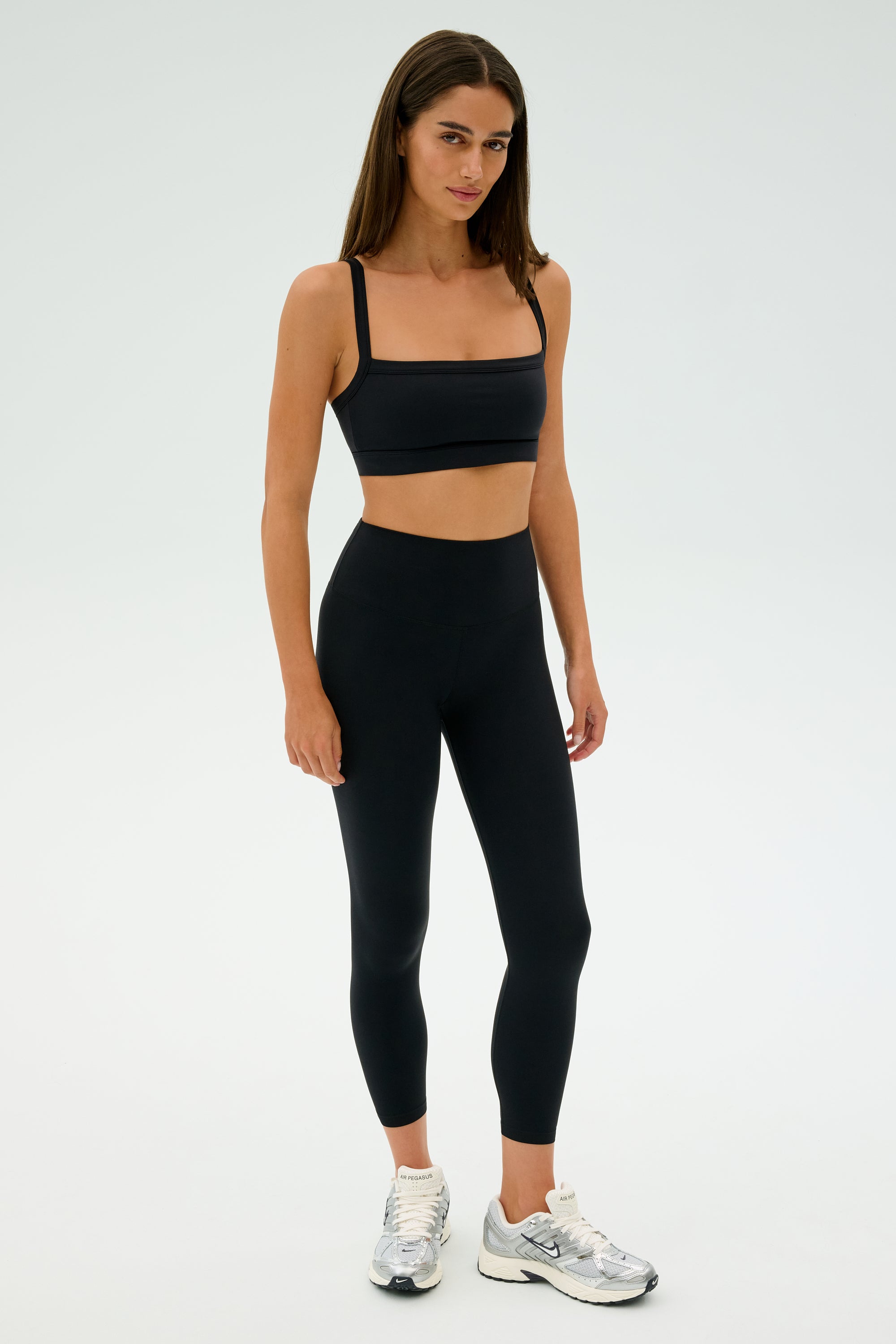 Amidst a minimalist setting, an individual stands confidently in a sleek black SPLITS59 Monah Rigor Bra paired with matching leggings and shiny silver sneakers—perfectly styled to conquer any gym workout with poise.