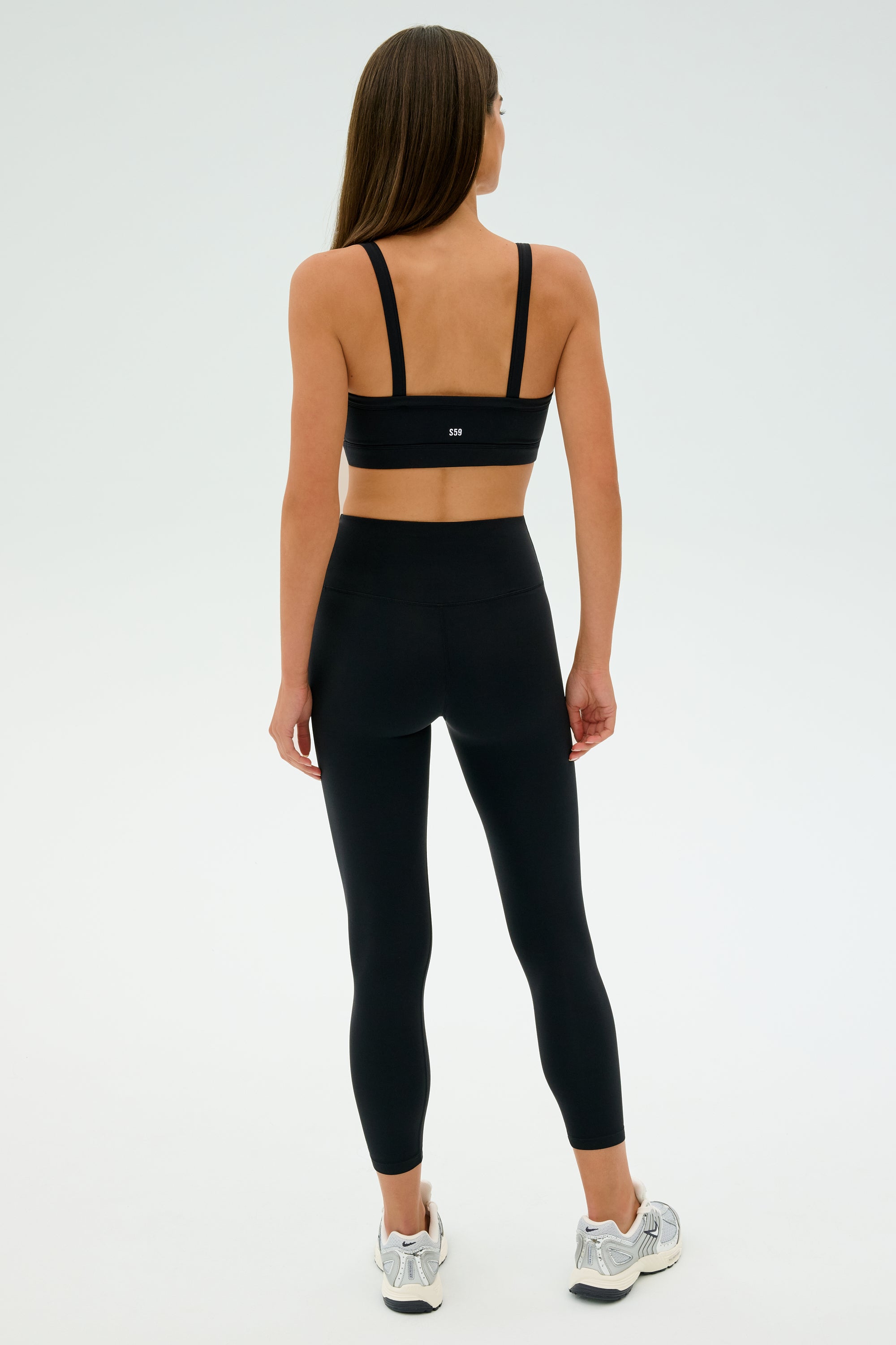 A woman models the SPLITS59 Monah Rigor Bra - Black, paired with black leggings and white sneakers, against a simple, light backdrop.