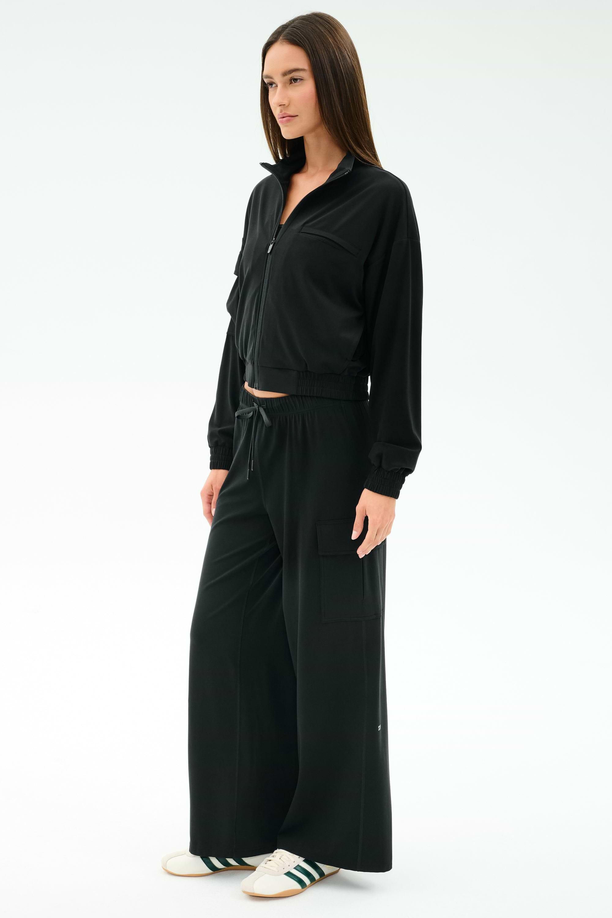 A woman stands against a plain white background wearing SPLITS59's black Alex Airweight Cargo Pant, styled with a matching black zip-up jacket and white sneakers featuring green stripes.