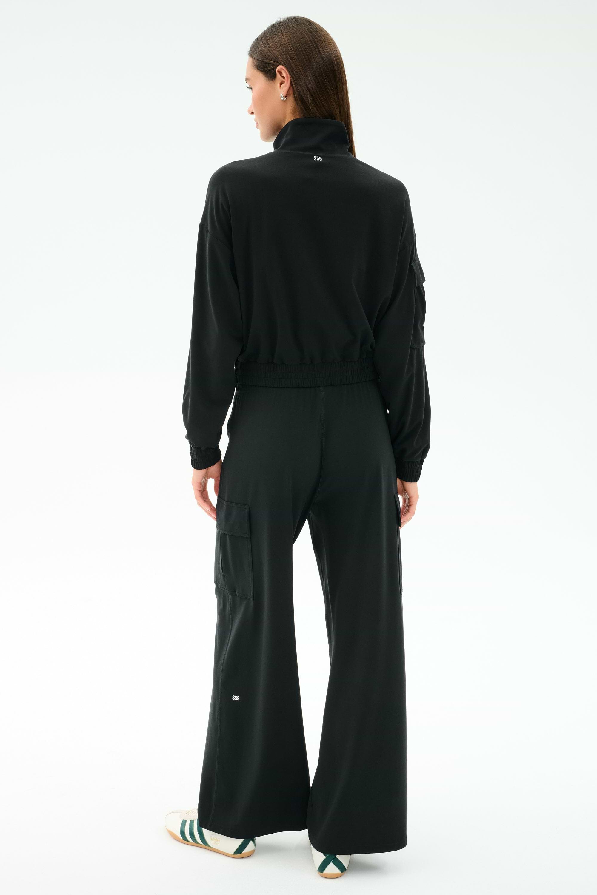 A woman stands facing away, wearing the SPLITS59 Alex Airweight Cargo Pant in black with white sneakers featuring green and gold stripes, paired with a black long-sleeve jacket against a plain white background.