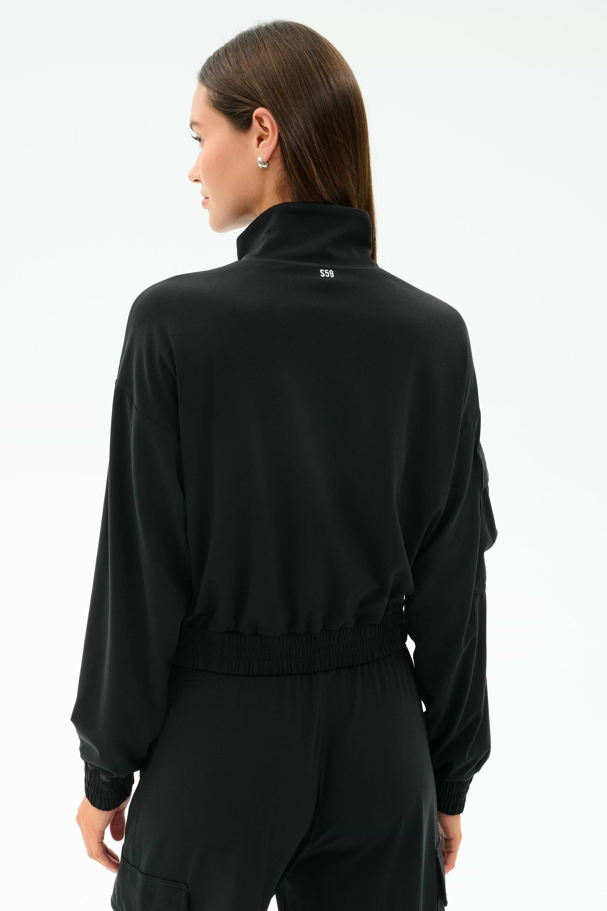 A woman with straight brown hair is seen from the back wearing the SPLITS59 Peaches Airweight Cargo Jacket in black against a plain light background.