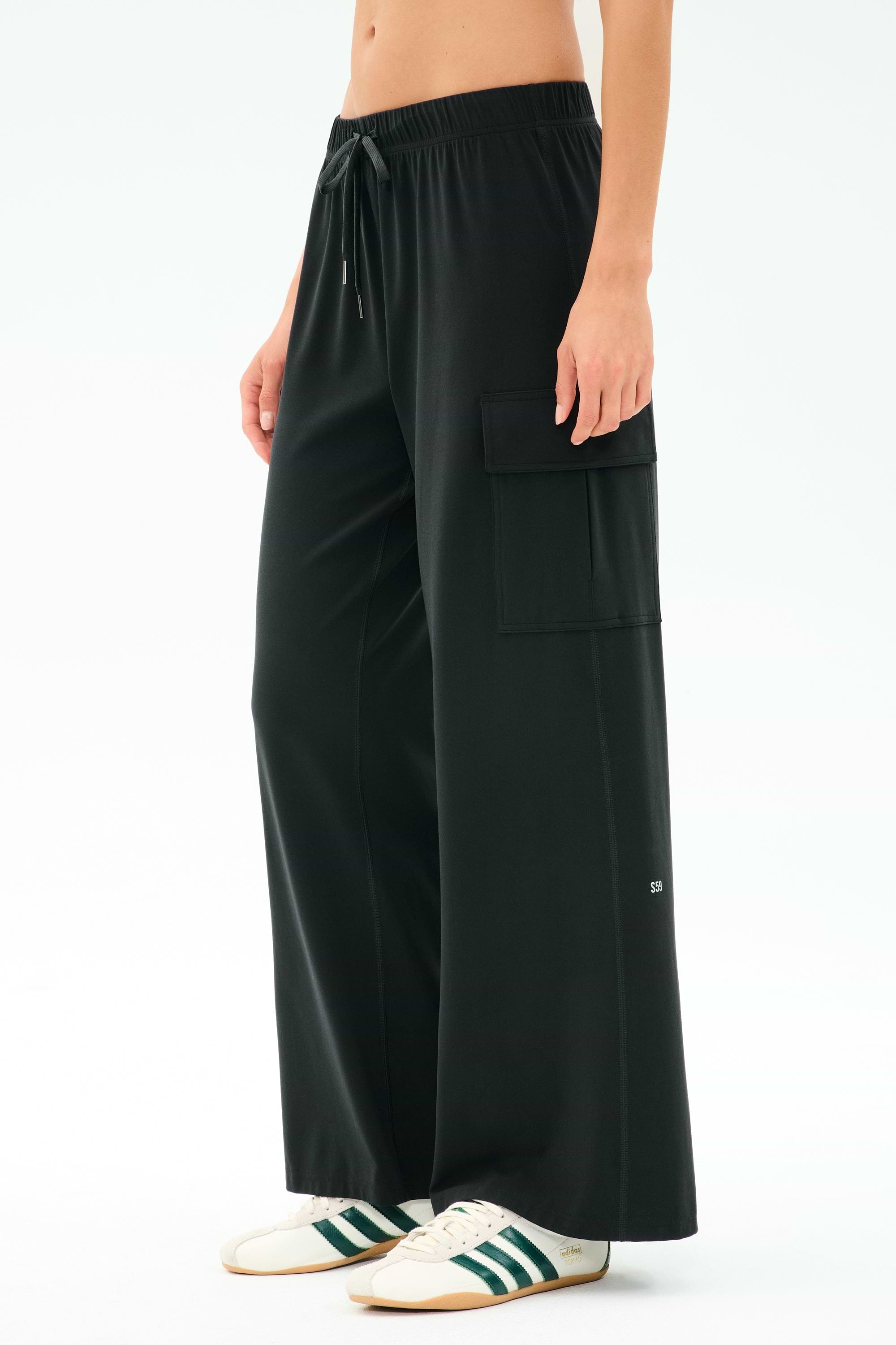A person wears the SPLITS59 Alex Airweight Cargo Pant in black, featuring a drawstring waist and side pocket, paired with white sneakers with green stripes.