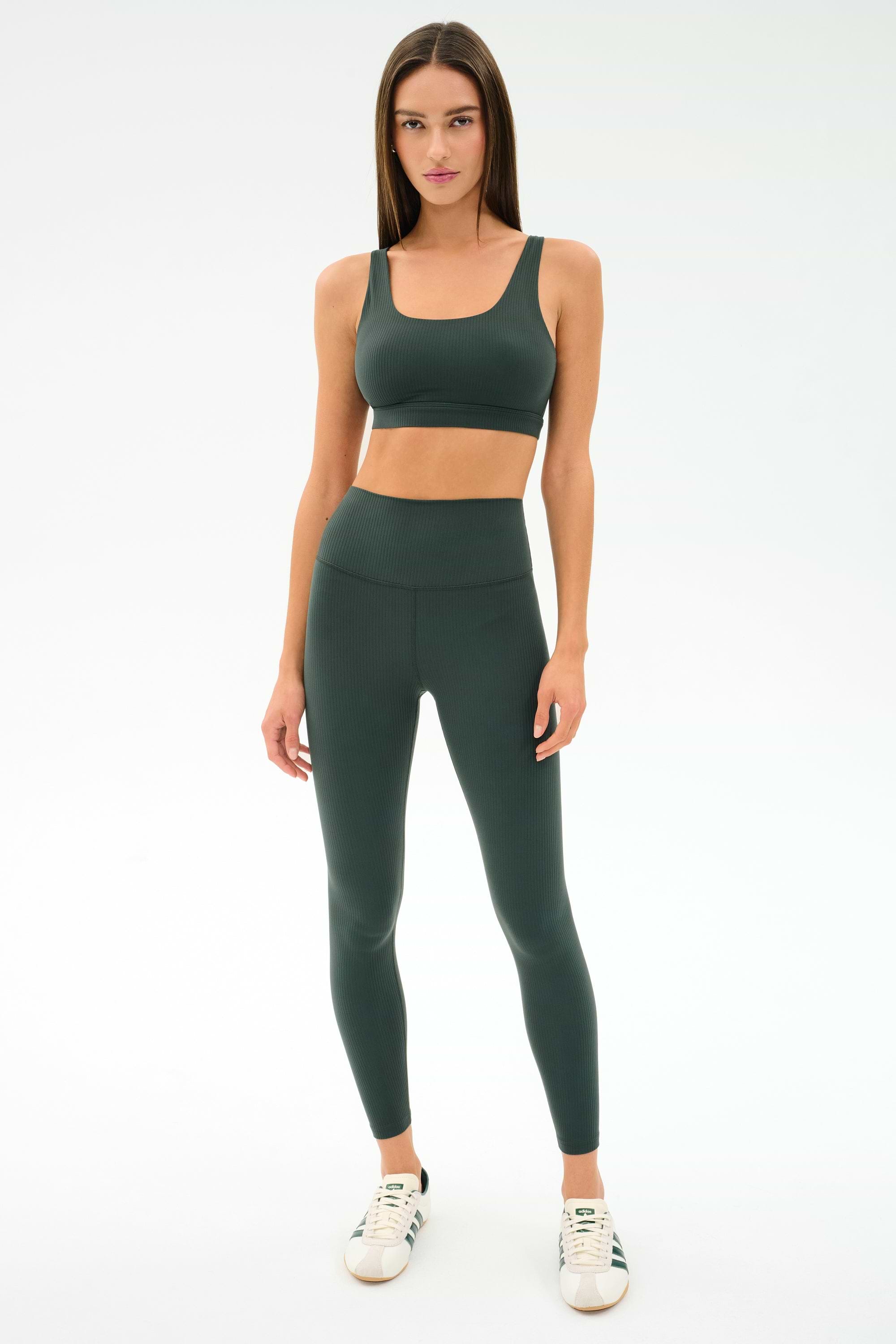 A woman stands against a white background wearing the SPLITS59 Airweight Rib Bra in Military, paired with matching leggings and white sneakers.