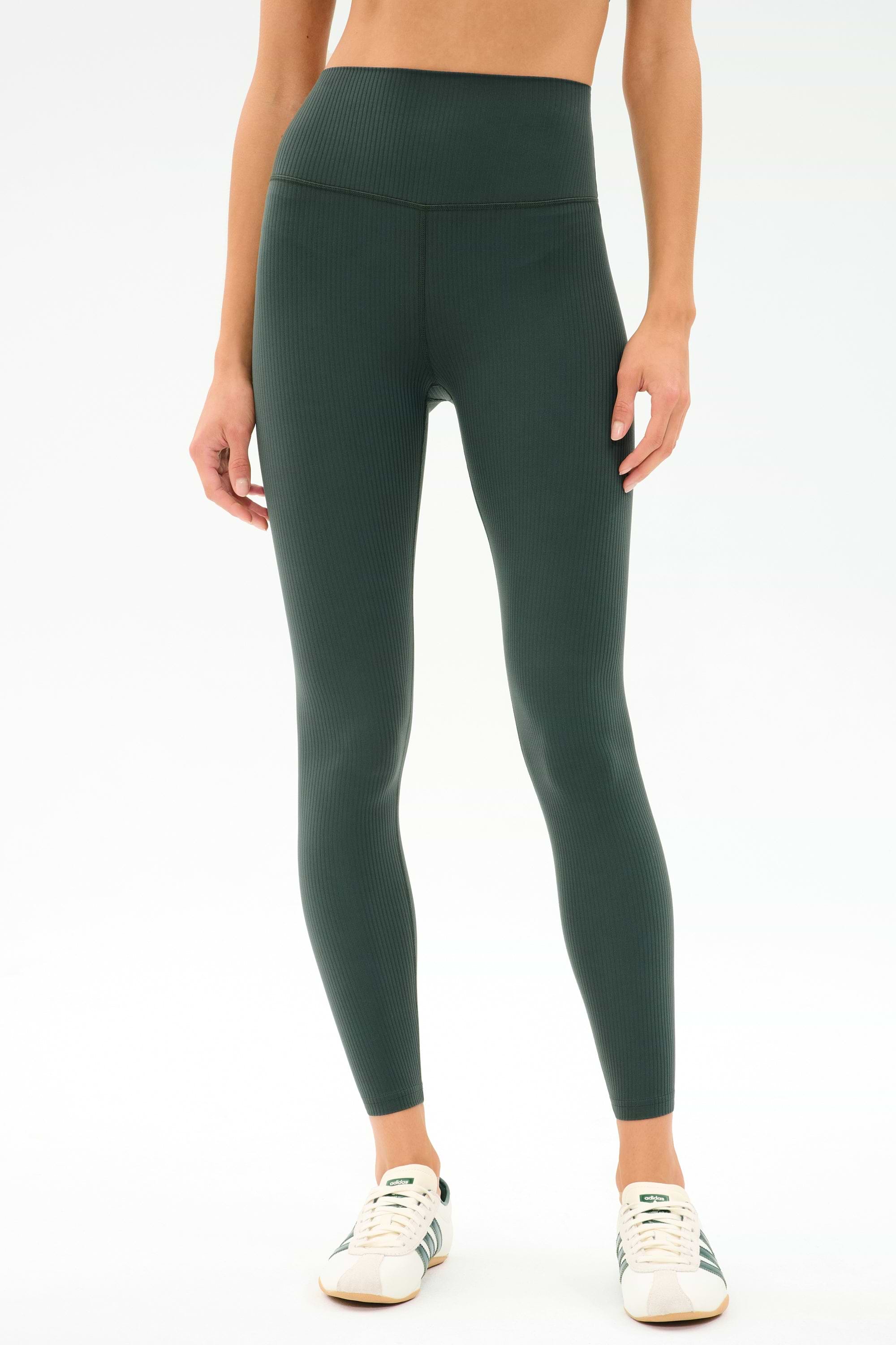 A person stands against a plain white background wearing SPLITS59 Airweight Rib High Waist 7/8 leggings in Military and white sneakers.