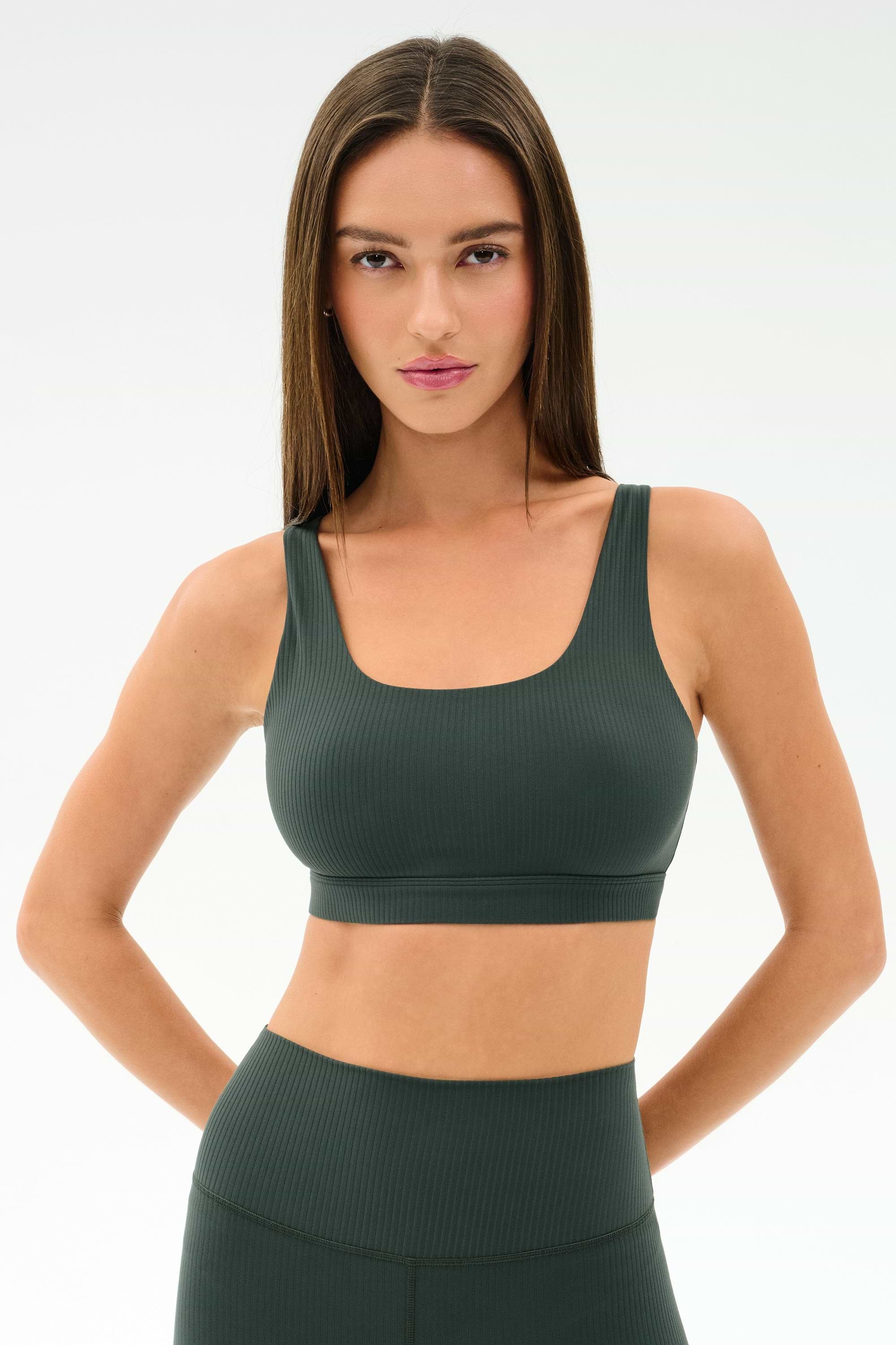 A woman with long brown hair wears the SPLITS59 Airweight Rib Bra in Military and matching high-waisted leggings, made from soft nylon spandex, standing with hands on hips against a plain white background.