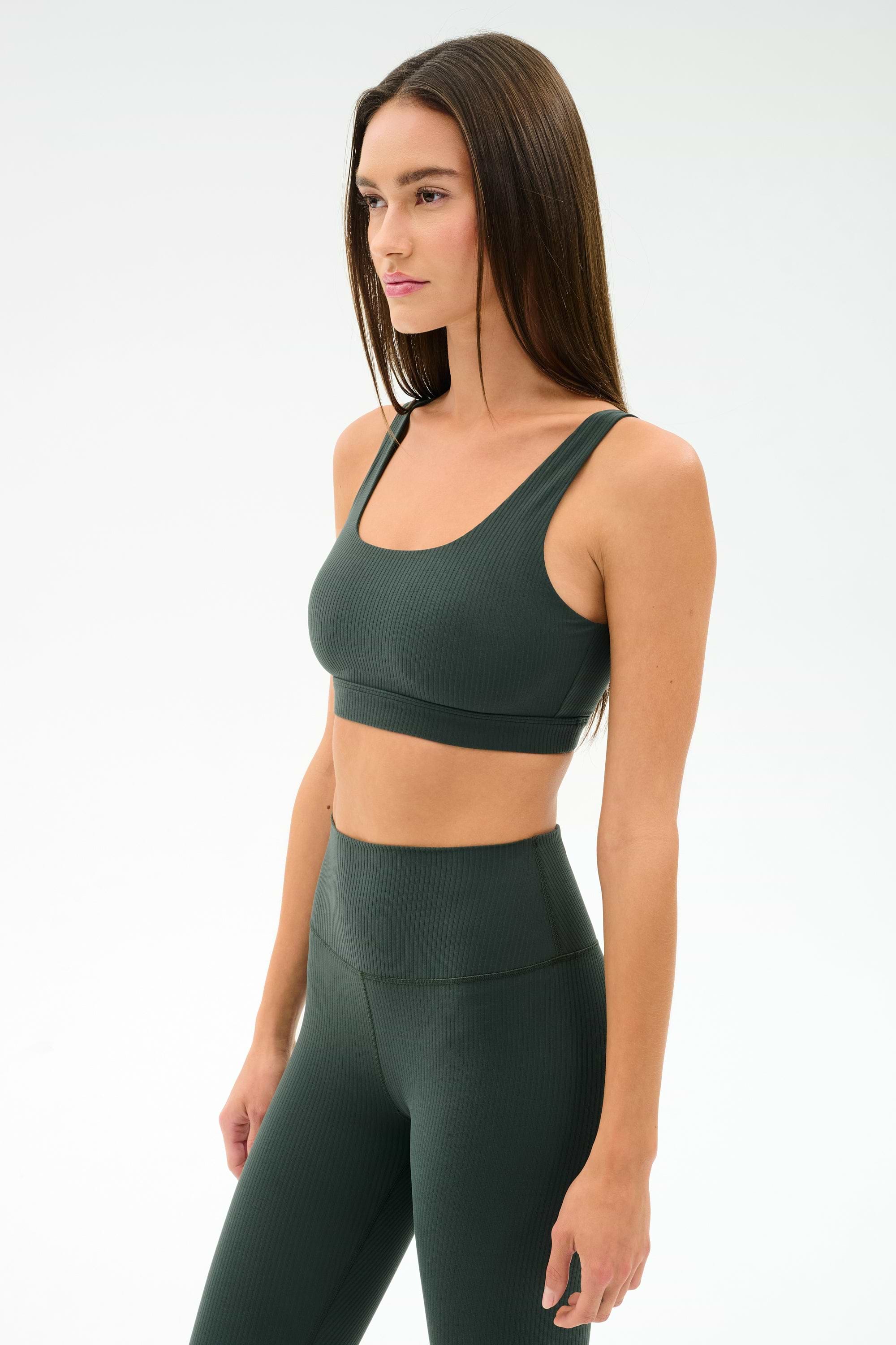 A woman wears the SPLITS59 Airweight Rib Bra in Military, paired with matching high-waisted leggings, both made from nylon spandex fabric, standing against a plain light background.