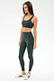 A woman poses against a plain background in the SPLITS59 Airweight Rib Bra - Military, a dark green nylon-spandex sports bra, paired with matching high-waisted leggings and white sneakers with green stripes.