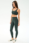 A woman poses against a plain background in the SPLITS59 Airweight Rib Bra - Military, a dark green nylon-spandex sports bra, paired with matching high-waisted leggings and white sneakers with green stripes.