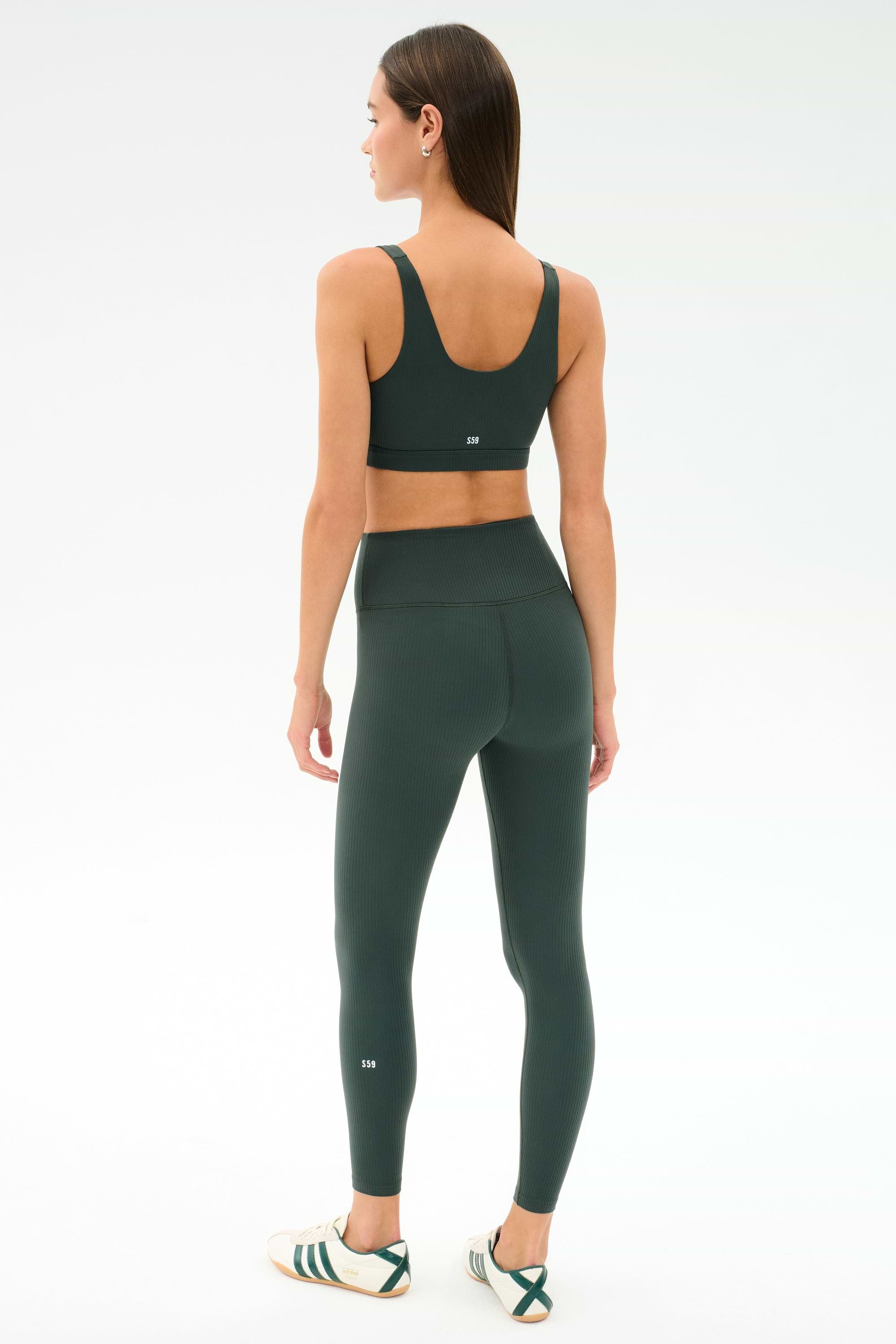 A woman faces away, wearing the SPLITS59 Airweight Rib Bra - Military, matching high-waisted leggings, and white sneakers with green stripes against a plain white background.
