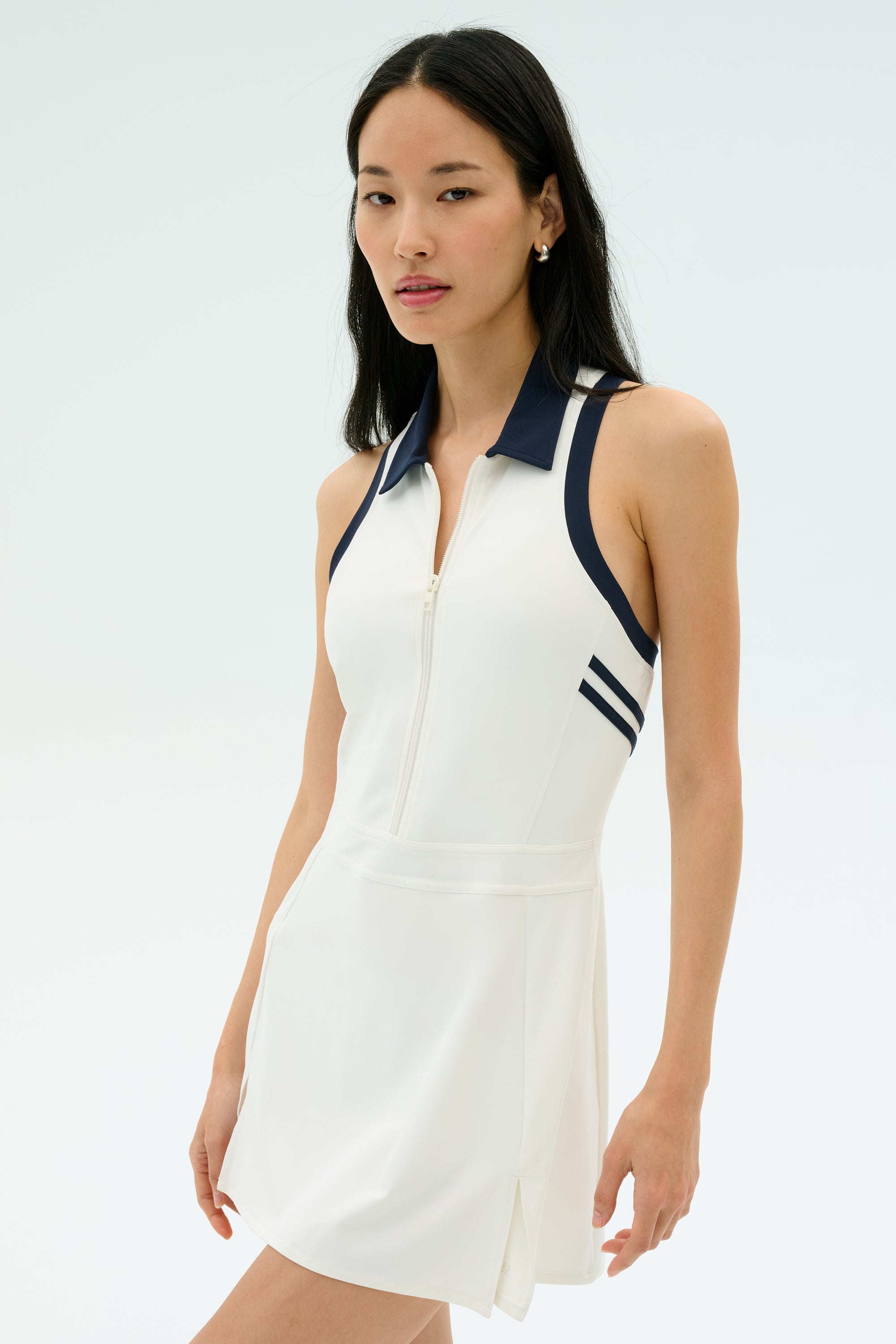 A woman wears the SPLITS59 Austin Rigor Dress in white/indigo, featuring a sleeveless design, zip-up front, and navy blue collar, standing against a plain light background.