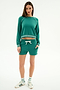 A woman stands against a white background wearing SPLITS59’s Reena Fleece Sweatshort in Arugula with a matching green long-sleeve top, white drawstring, striped socks, and white sneakers.