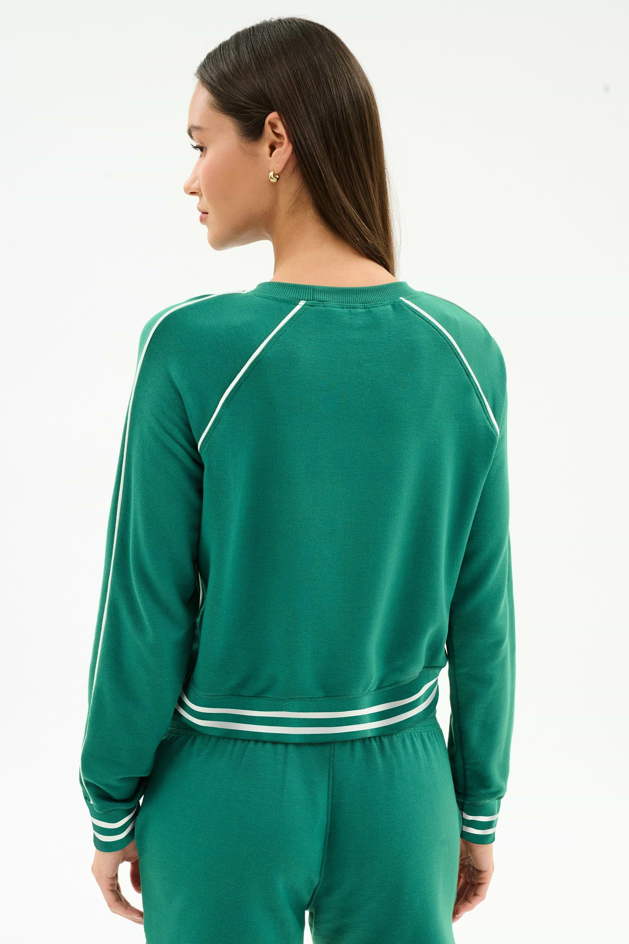 A woman with long straight hair and gold stud earrings wears the SPLITS59 Brooks Fleece Sweatshirt With Stripe in Arugula/White, featuring soft modal fabric and white stripes on the sleeves, cuffs, and waist.