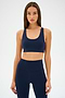 An individual in an Ella Airweight Bra by SPLITS59, in an Indigo/Black Currant color scheme, stands against a plain backdrop, highlighting its sleek racer back design.