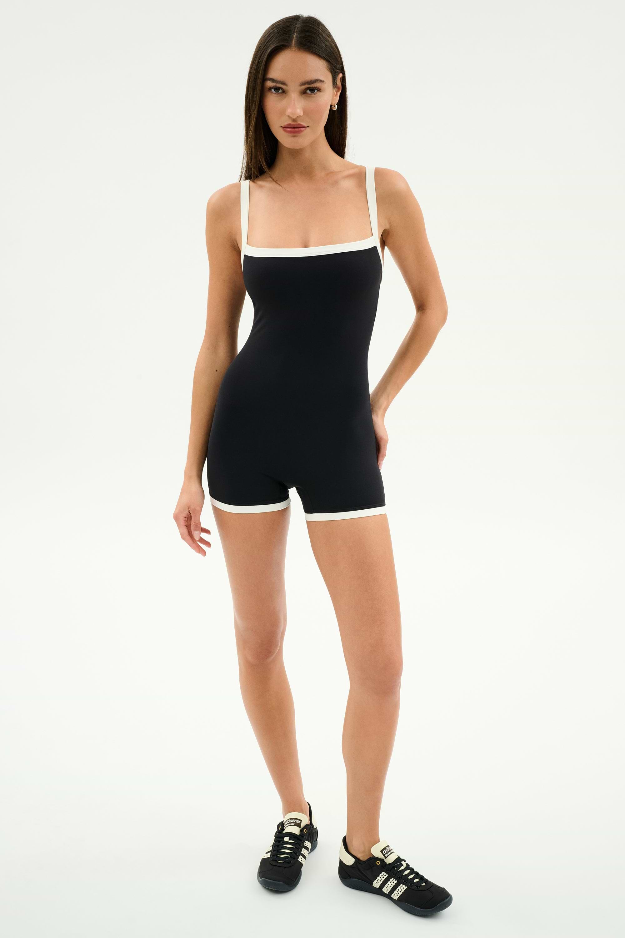 A woman stands against a plain background wearing the SPLITS59 Monah Rigor Jumpsuit in black/white—made from supportive Rigor fabric for high-impact workouts—and black sneakers with white stripes.