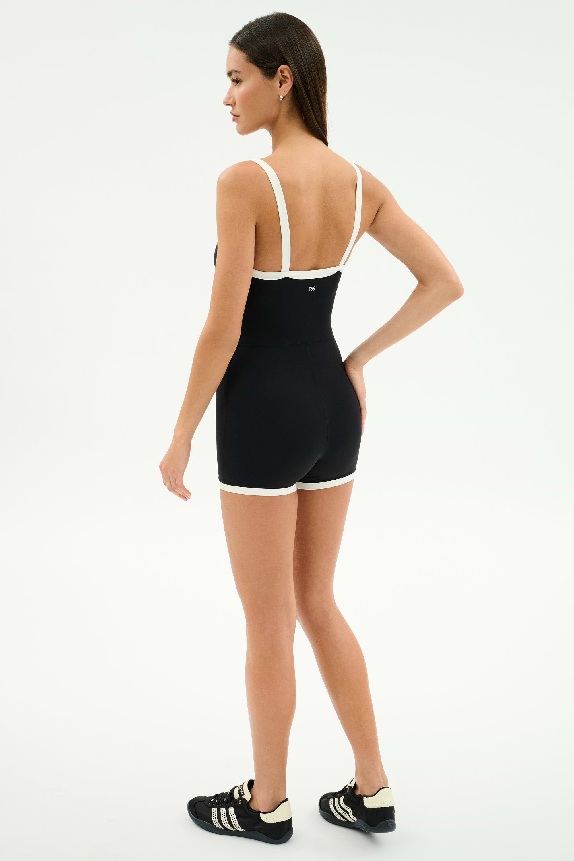 A woman faces away, wearing the SPLITS59 Monah Rigor Jumpsuit in Black/White with black Adidas sneakers, against a plain white background.