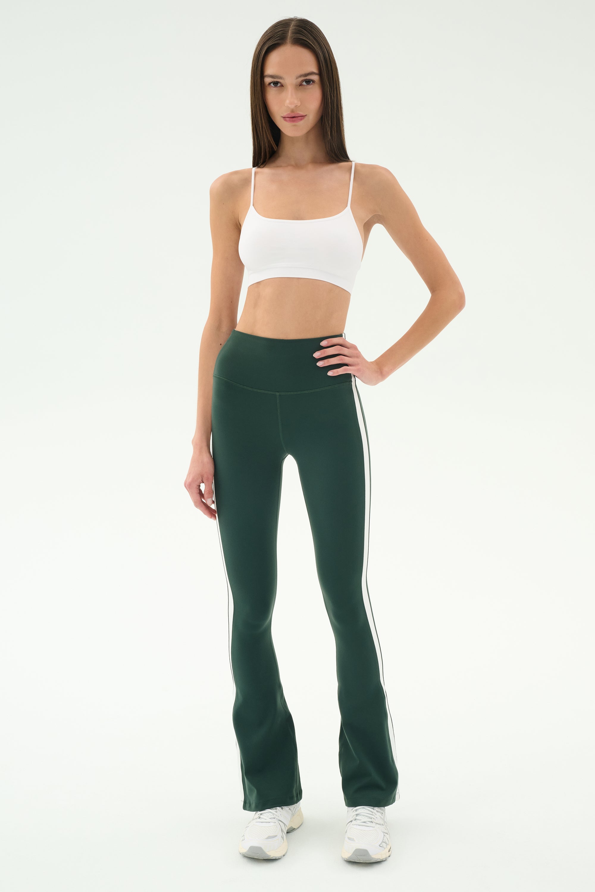 A woman wears the SPLITS59 Raquel High Waist Flared Legging in Military/White, paired with a white crop top and white sneakers, standing against a plain white background.