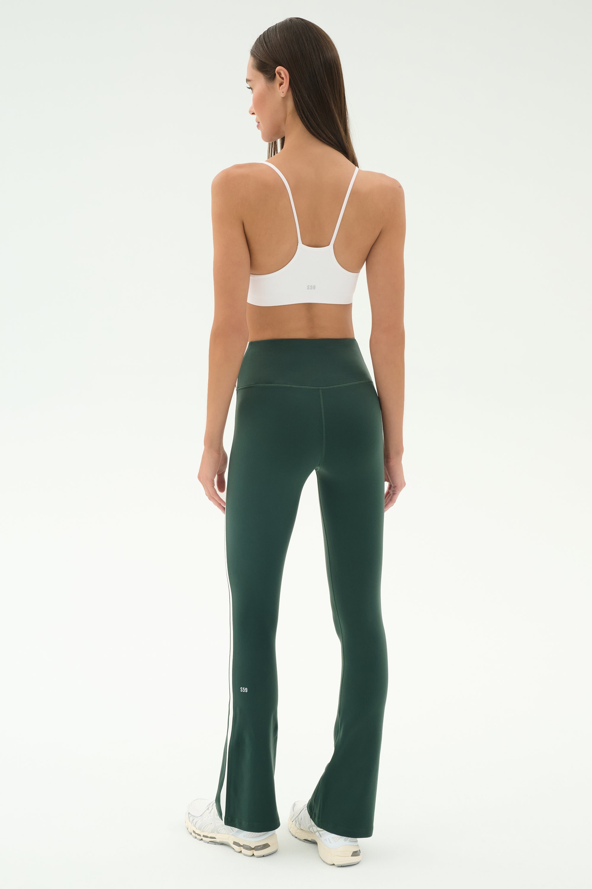 A woman faces away, wearing SPLITS59's Raquel High Waist Flared Legging in Military/White and a white sports bra, paired with white sneakers against a plain light background.