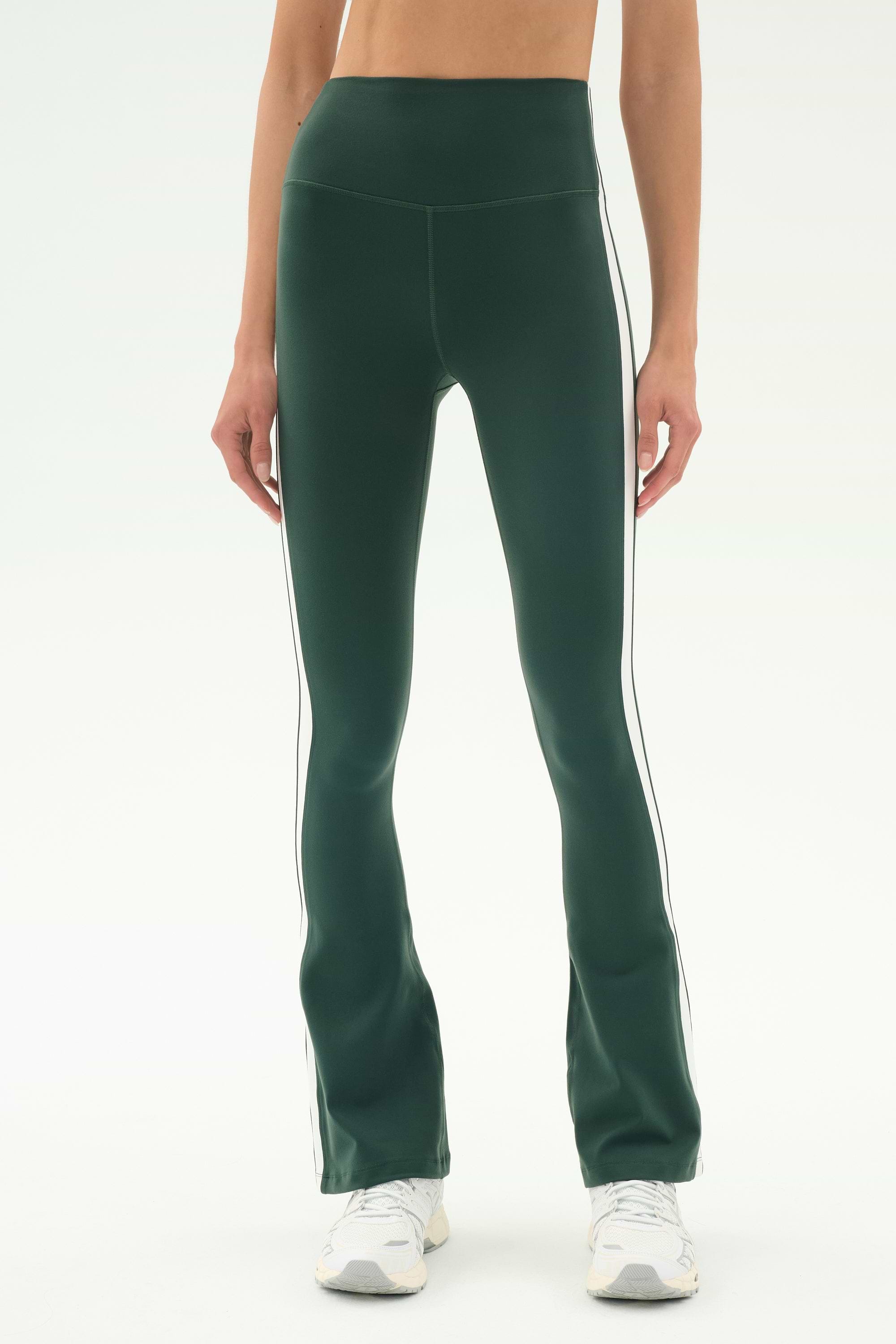 A person wears SPLITS59's Raquel High Waist Flared Legging in Military/White with white piping, paired with white sneakers, standing against a plain white background.