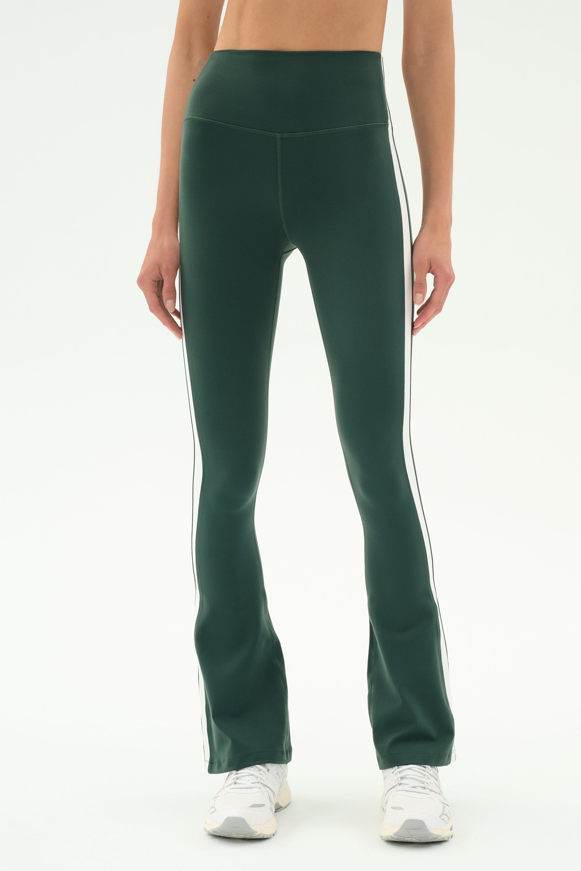 A person wears SPLITS59's Raquel High Waist Flared Legging in Military/White with white piping, paired with white sneakers, standing against a plain white background.