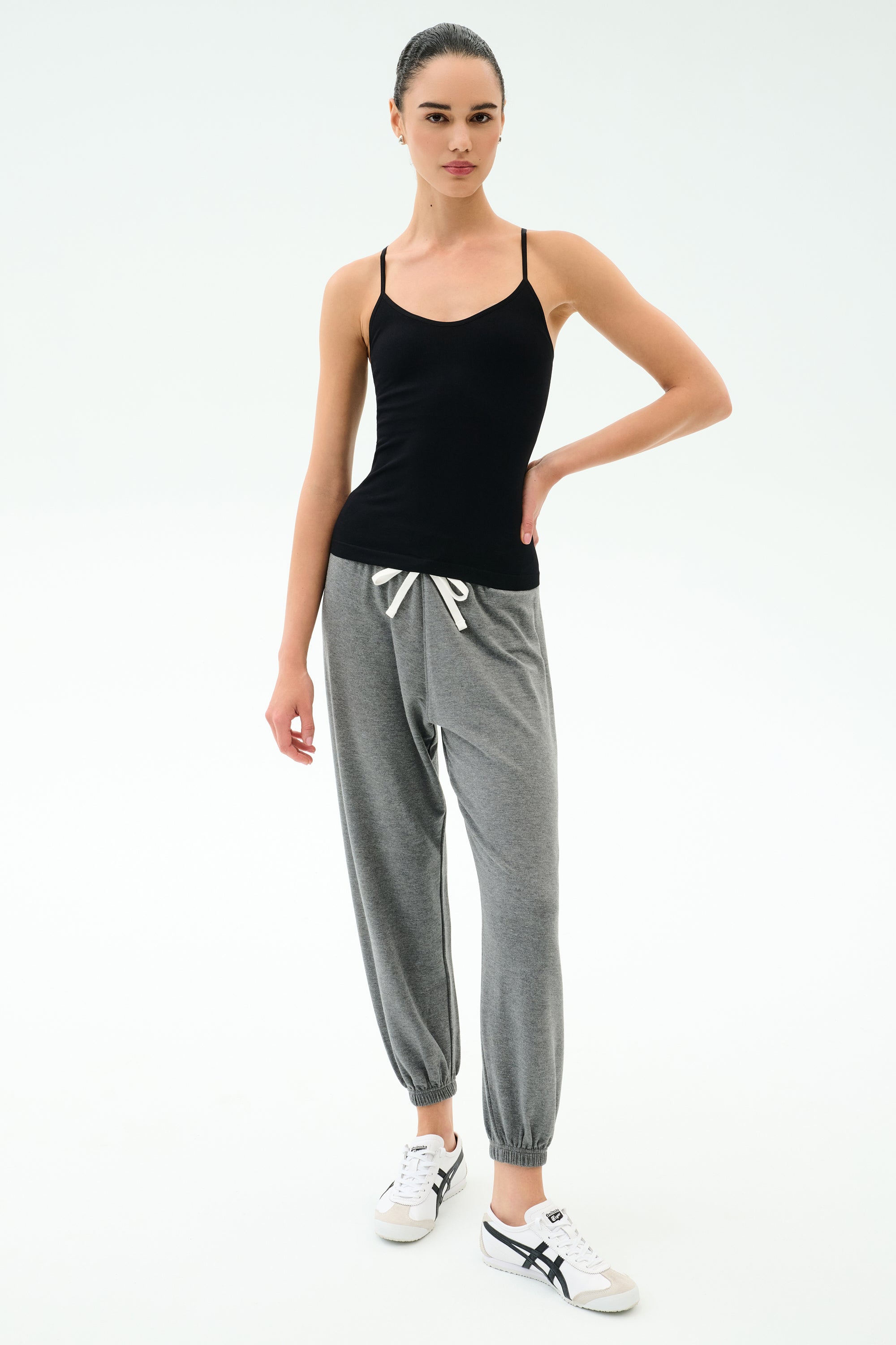 A woman stands against a white background wearing the SPLITS59 Loren Seamless Waist Length Tank in black, paired with gray drawstring jogger pants and white sneakers.