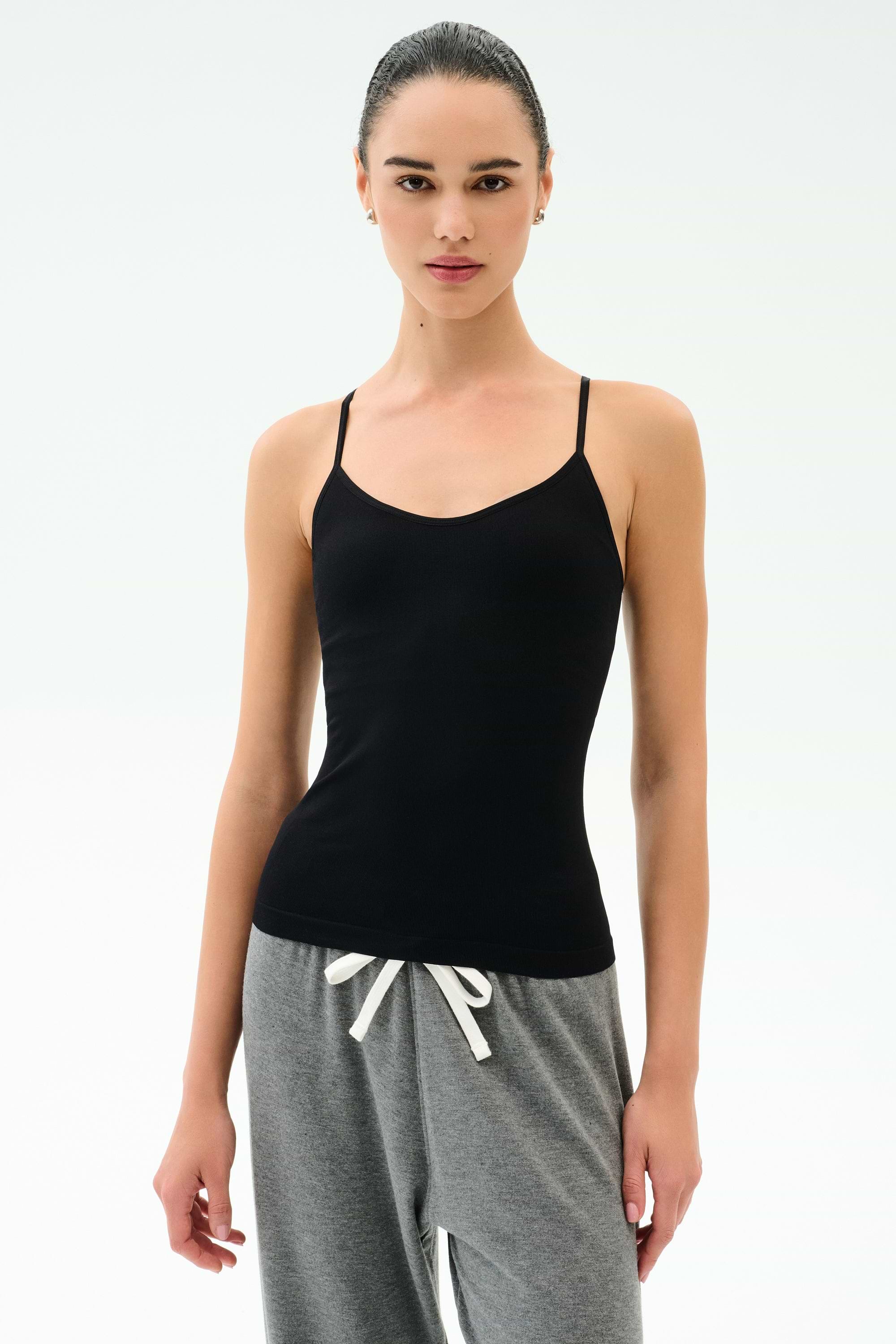 A person wears the SPLITS59 Loren Seamless Waist Length Tank in black, featuring a built-in shelf bra, paired with gray drawstring pants, standing against a plain white background.