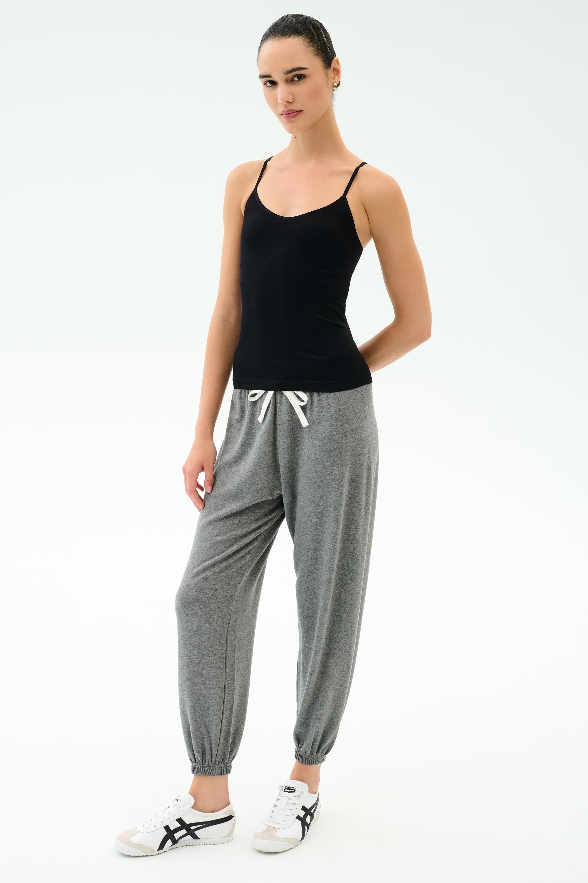 A person stands against a plain background wearing the SPLITS59 Loren Seamless Waist Length Tank in black, paired with gray drawstring sweatpants and white sneakers.