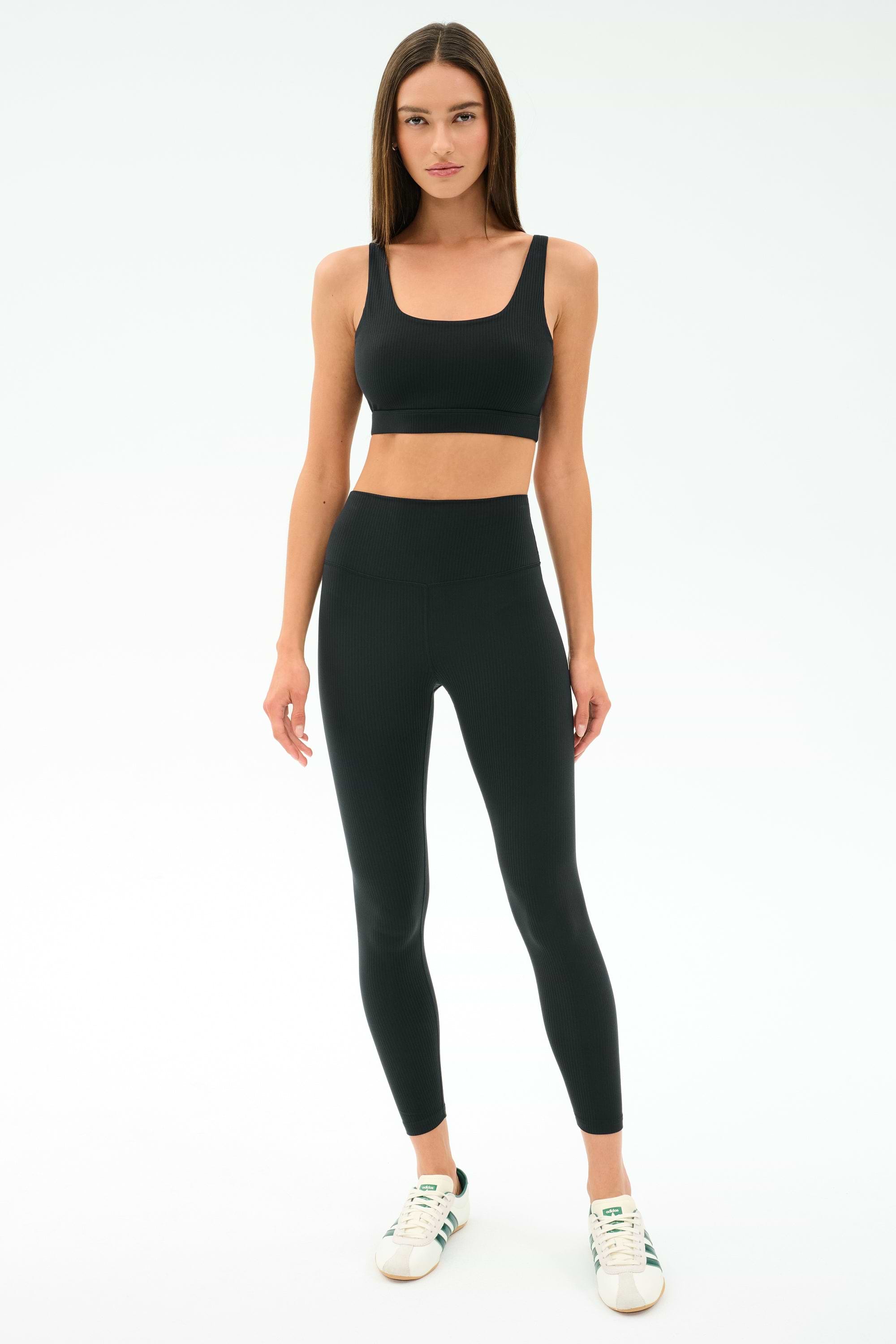 A woman stands facing forward, wearing the SPLITS59 Airweight Rib High Waist 7/8 leggings in black with a matching sports bra and white sneakers against a plain white background—ideal for Pilates or hot yoga.