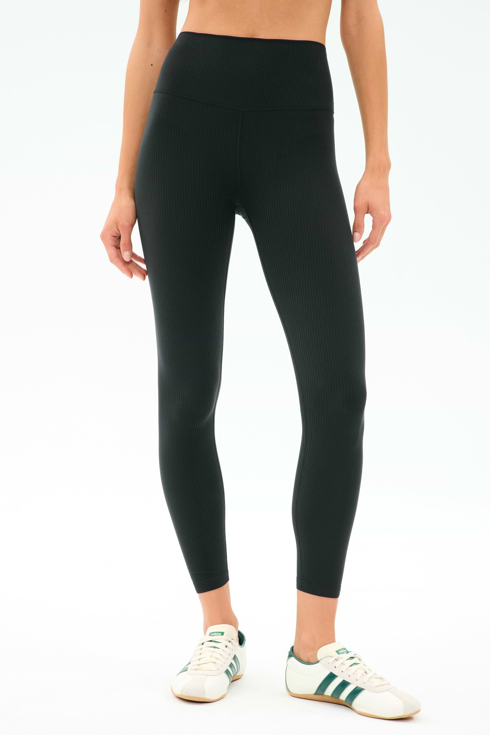 A person wears SPLITS59 Airweight Rib High Waist 7/8 leggings in black—ideal for Pilates or hot yoga—with white sneakers featuring green stripes, standing against a simple light background.