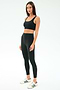 A woman faces forward wearing SPLITS59’s Airweight Rib High Waist 7/8 leggings in black with a matching sports bra and white sneakers with green stripes, set against a plain white background—ideal for Pilates or hot yoga.