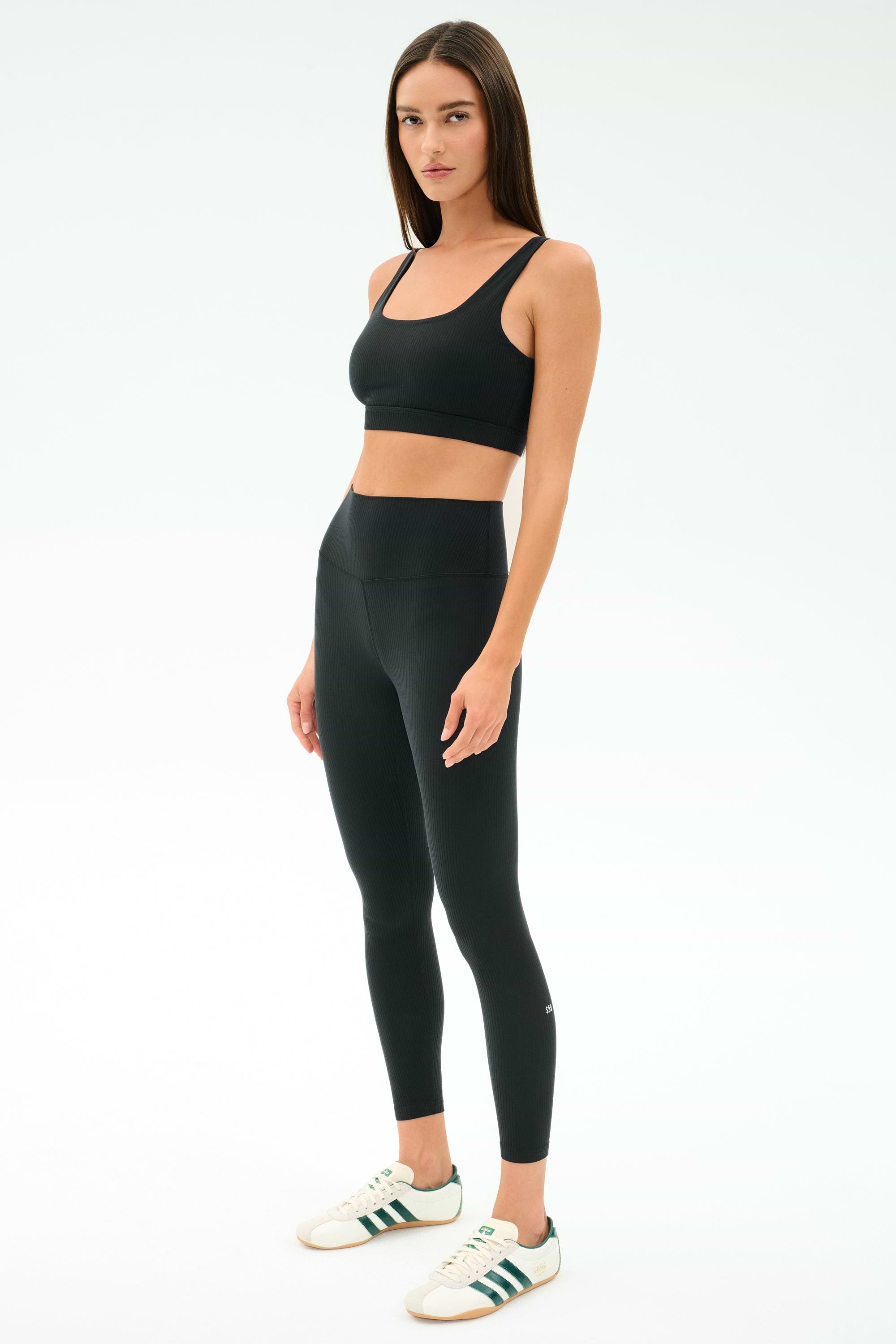 A woman faces forward wearing SPLITS59’s Airweight Rib High Waist 7/8 leggings in black with a matching sports bra and white sneakers with green stripes, set against a plain white background—ideal for Pilates or hot yoga.