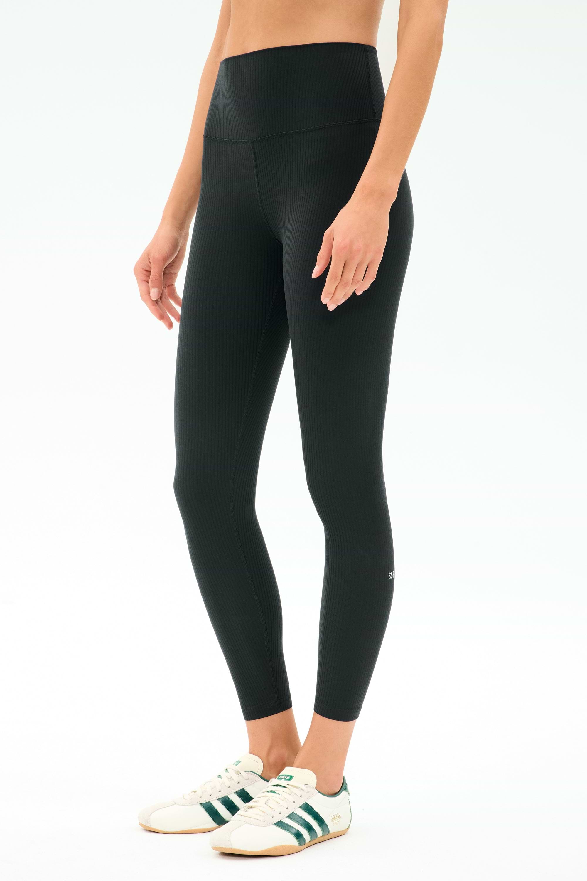 A person wears SPLITS59's Airweight Rib High Waist 7/8 leggings in black, ideal for Pilates or hot yoga, paired with white sneakers featuring green stripes, standing against a plain light background.