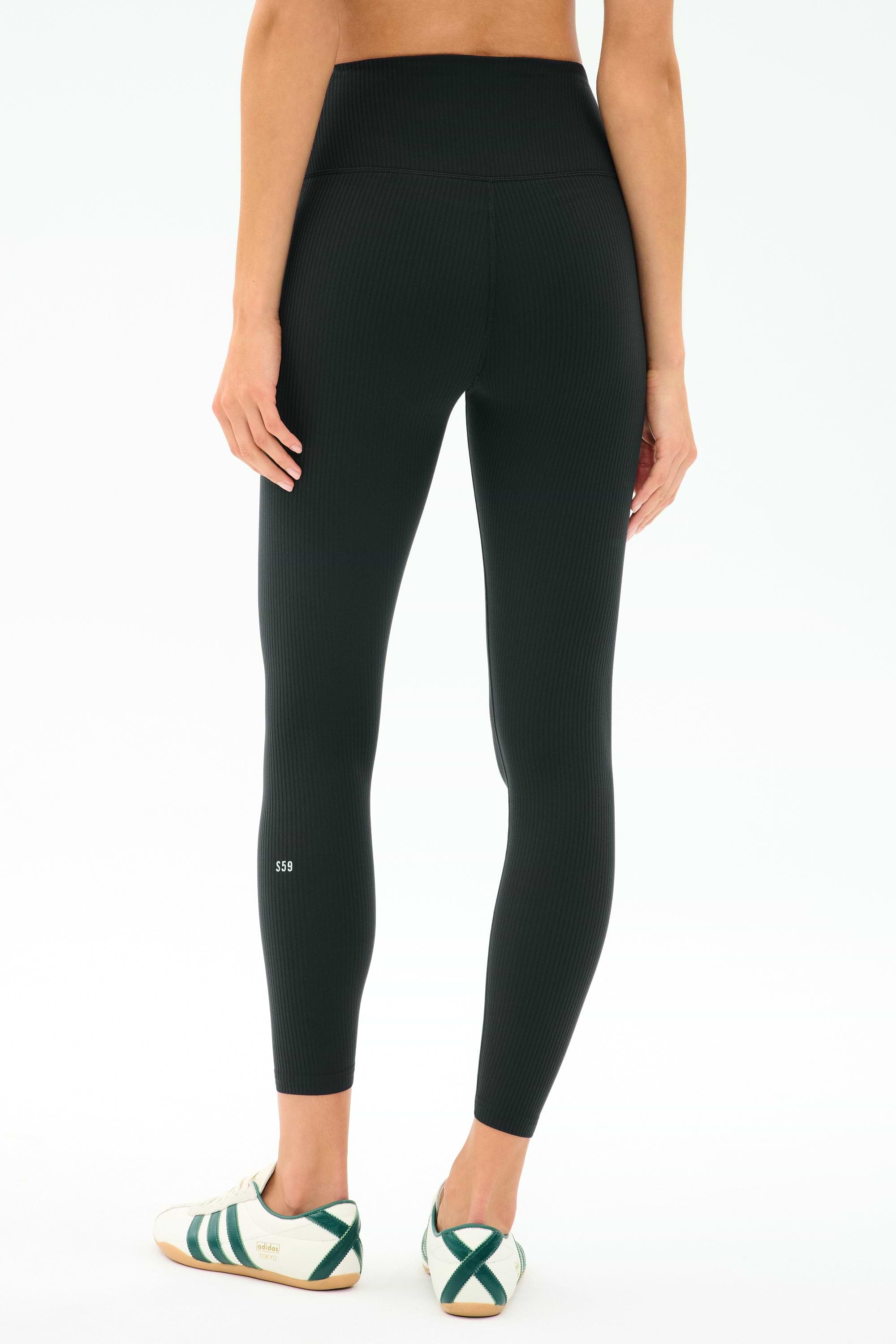 Wearing SPLITS59's Airweight Rib High Waist 7/8 leggings in black, a person stands with their back to the camera in white athletic shoes with green stripes, ready for hot yoga or Pilates against a plain background.